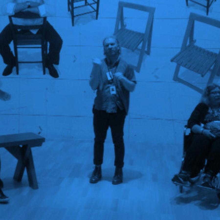 Blue-toned photo of a guide wearing a lanyard, standing in a gallery and talking with a group seated on benches, one in a wheelchair. Wooden chairs are mounted on the wall behind them.