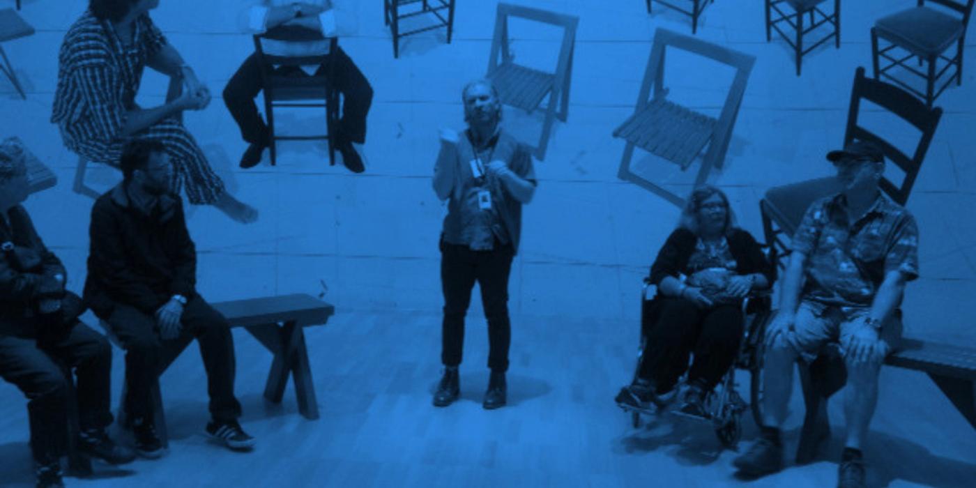 Blue-toned photo of a guide wearing a lanyard, standing in a gallery and talking with a group seated on benches, one in a wheelchair. Wooden chairs are mounted on the wall behind them.