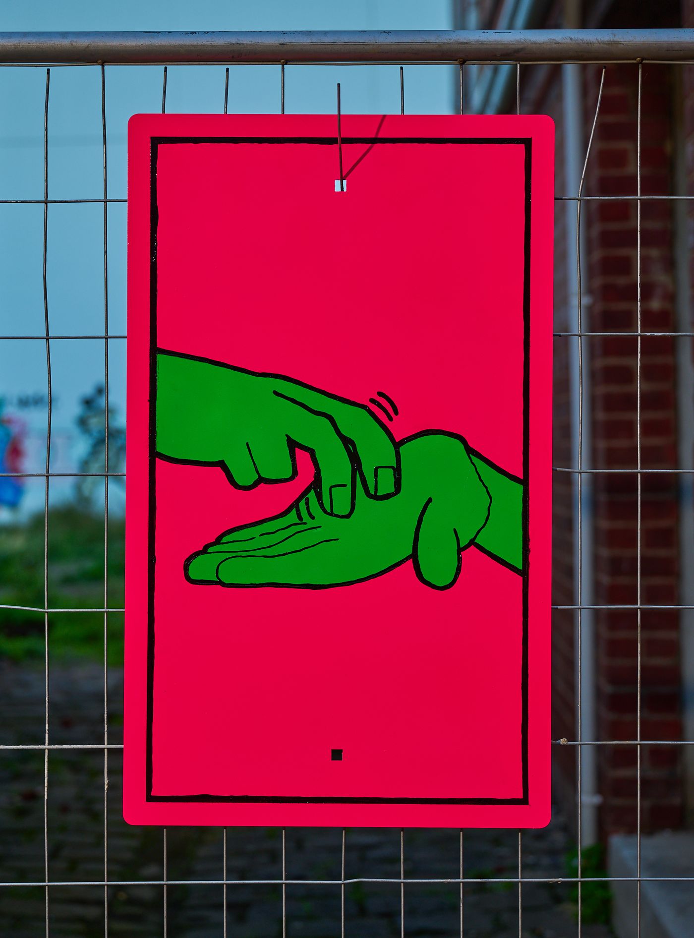 Series plate — two green hands signing on fluoro pink, hung on wire fencing
