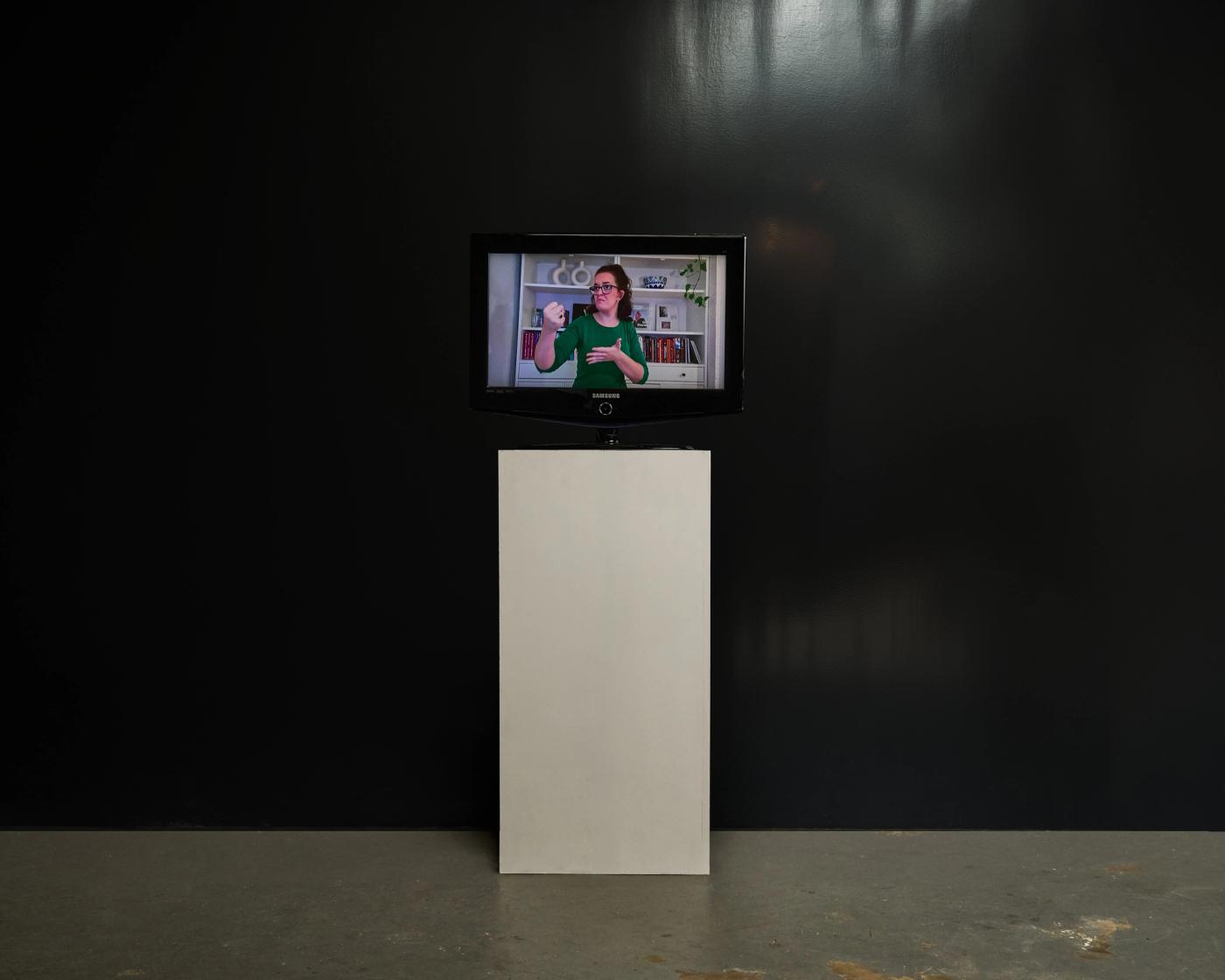 Breaking the fault lines — a monitor on a white plinth against a black wall, playing a signed video