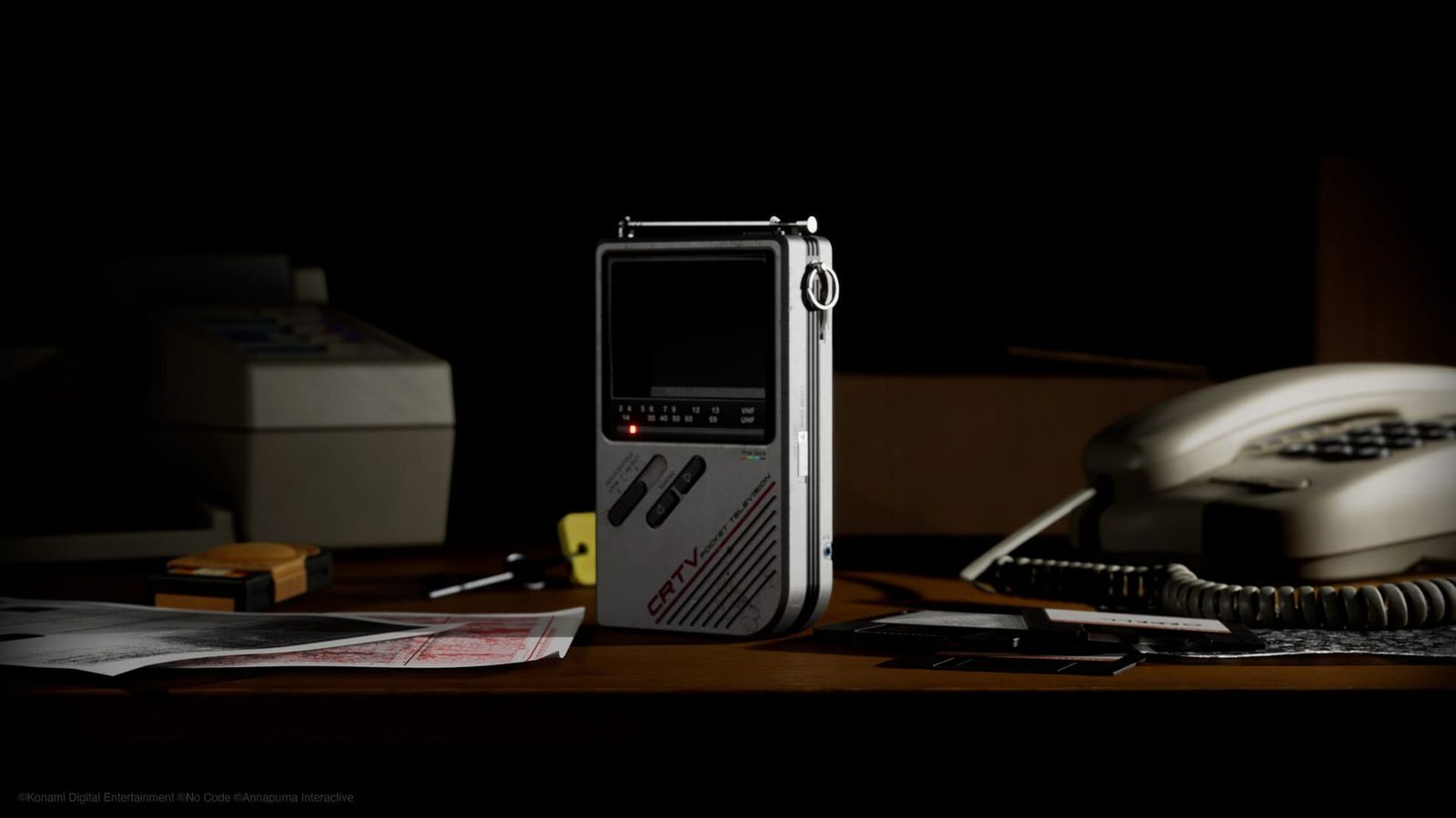 A grey handheld CRT television device on a dark desk next to a telephone and papers