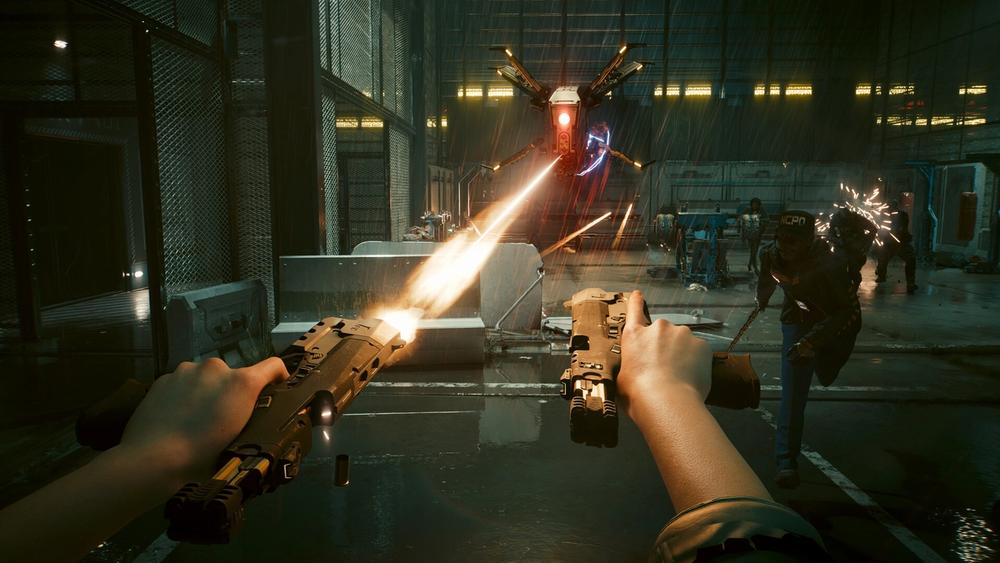 First-person view of V firing two pistols at once inside a warehouse while a drone hovers overhead in Cyberpunk 2077