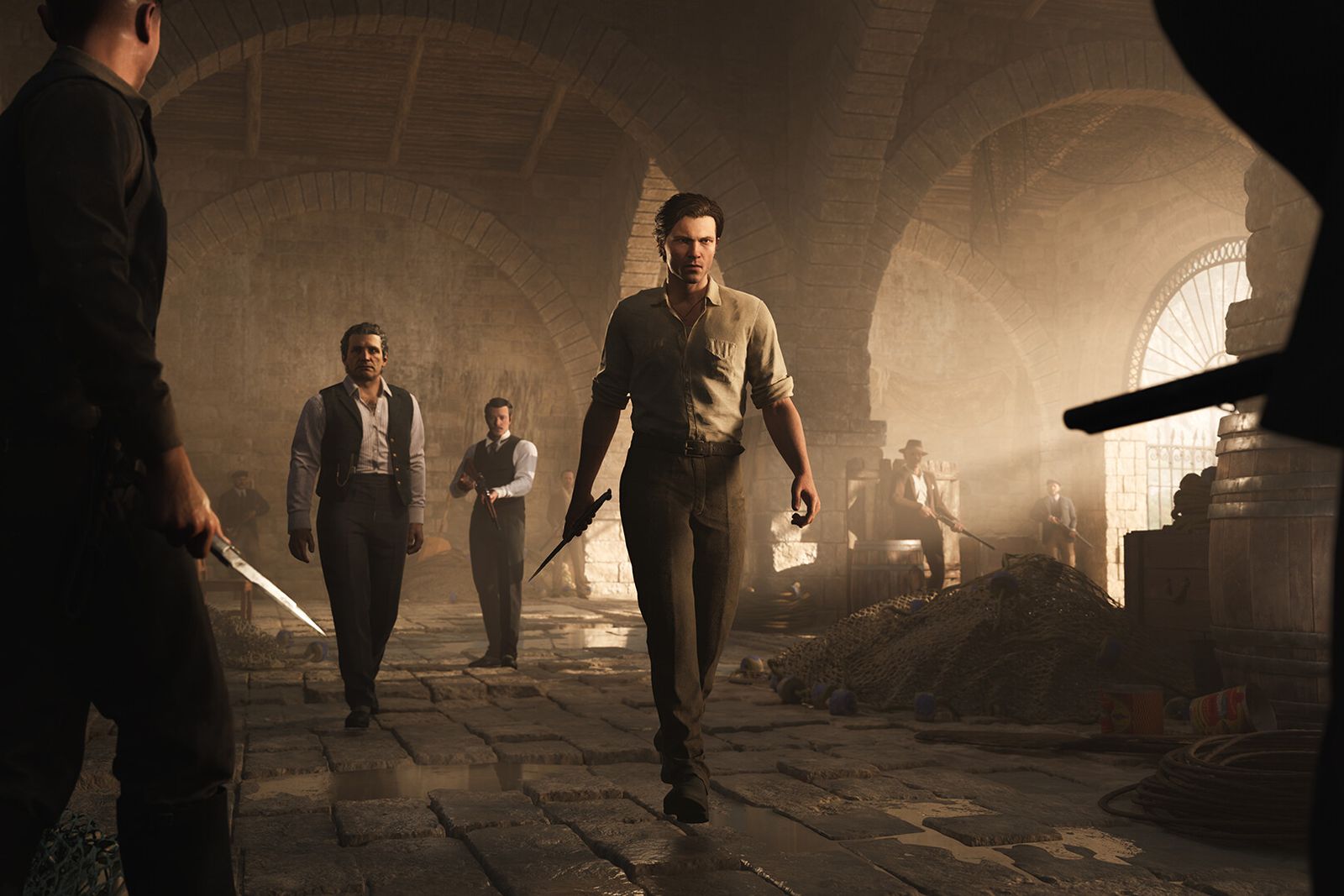 Men in early 1900s clothing, one holding a knife, face off in a dusty stone warehouse in Mafia: The Old Country