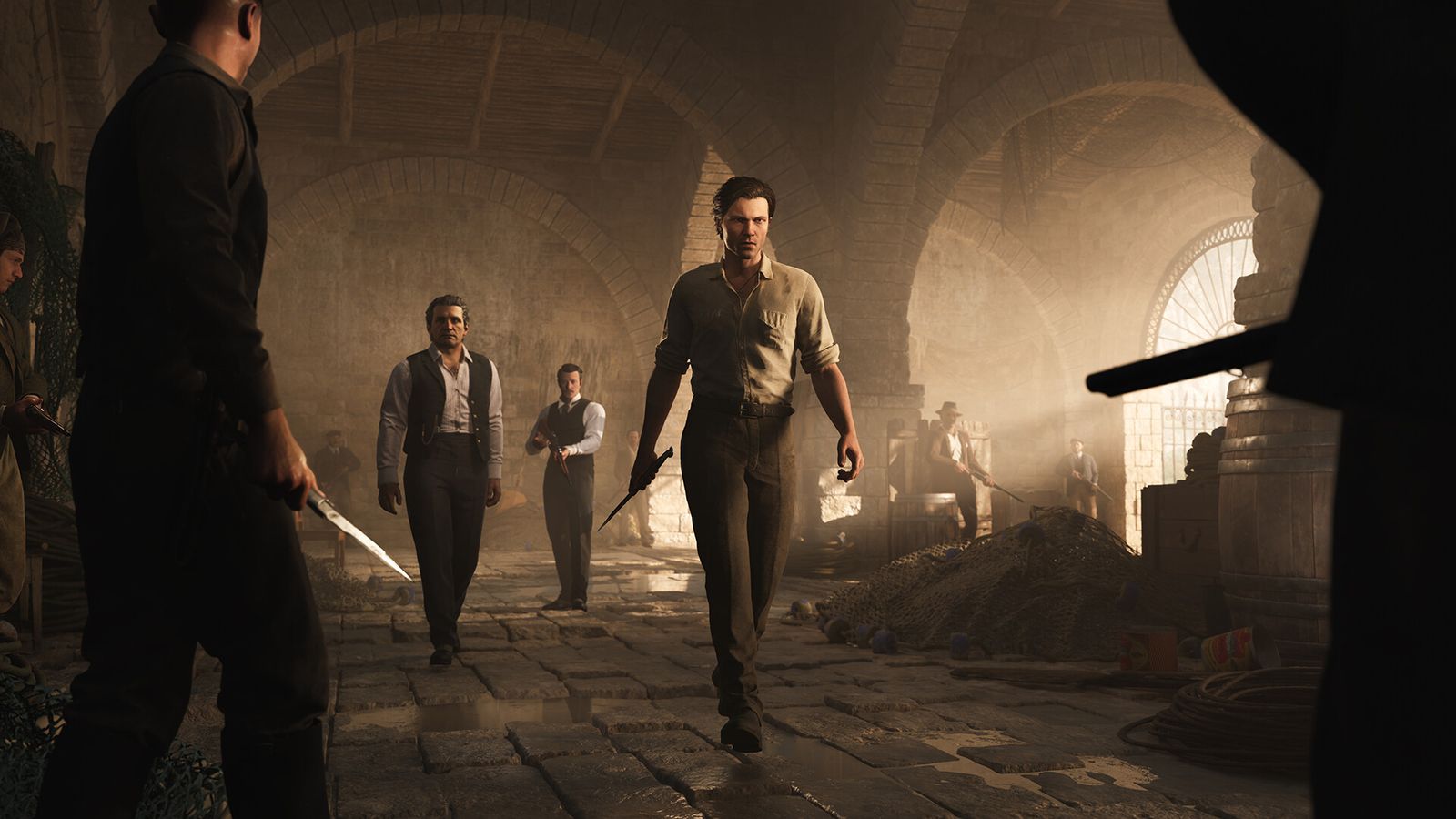 Men in early 1900s clothing, one holding a knife, face off in a dusty stone warehouse in Mafia: The Old Country
