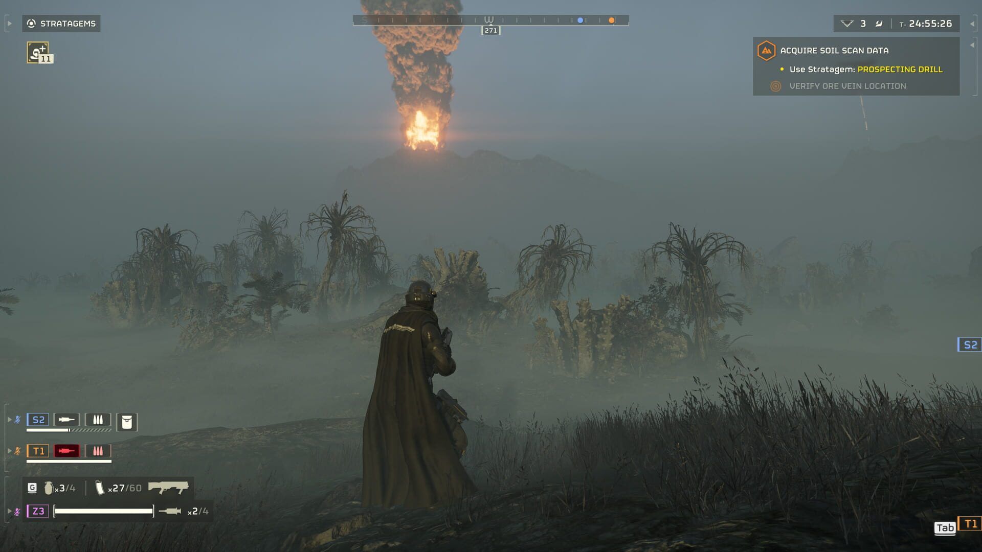 A Helldiver in a long coat stands in foggy jungle grass as a column of fire and smoke rises in the distance.