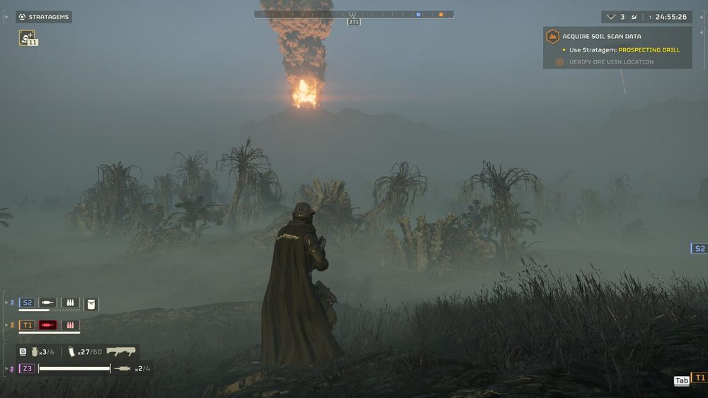 A Helldiver in a long coat stands in foggy jungle grass as a column of fire and smoke rises in the distance.