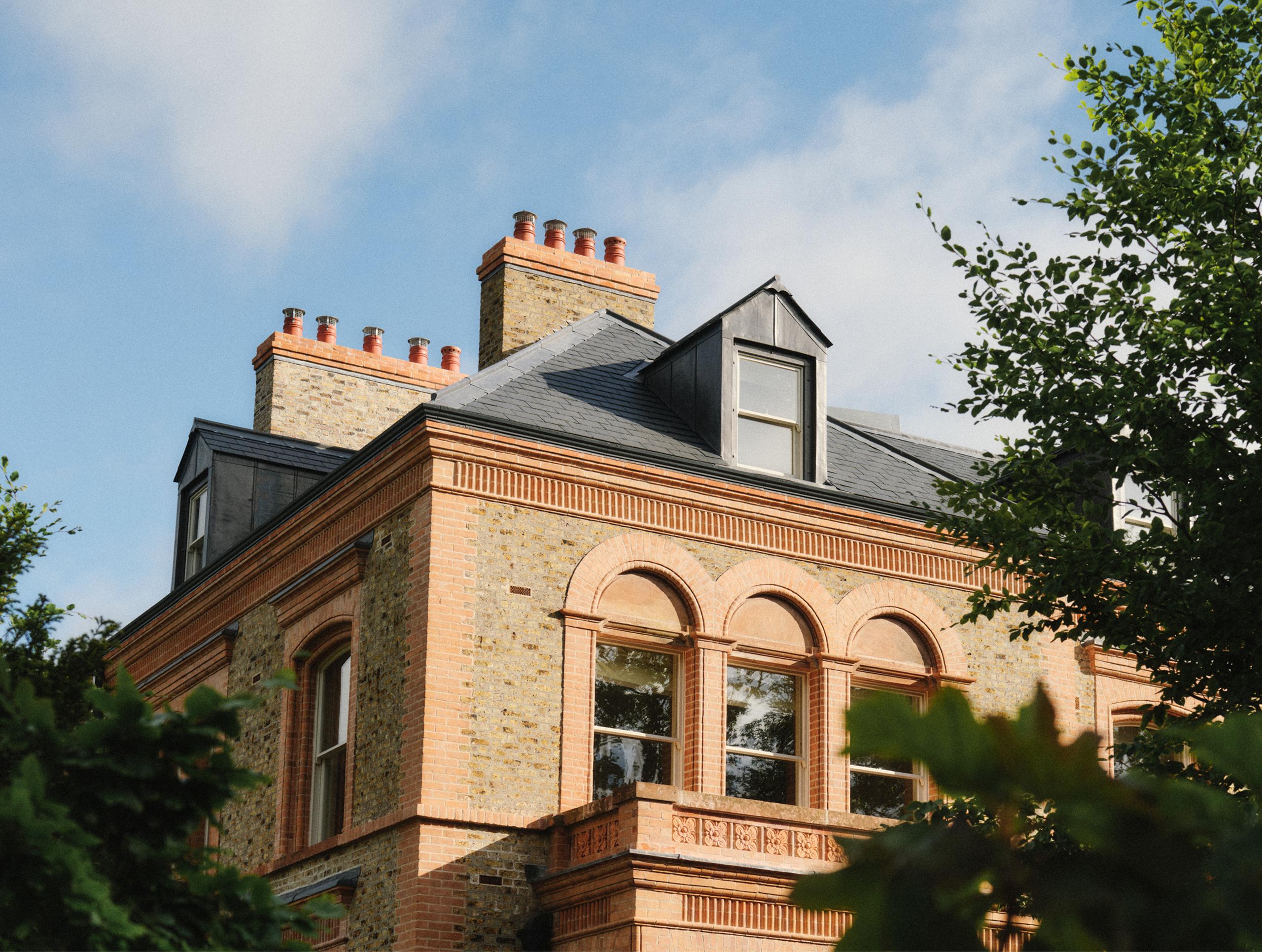 Honey-coloured stone Victorian guesthouse with slate roof, brick-trimmed arched windows and tall chimneys, framed by green summer foliage against a blue sky