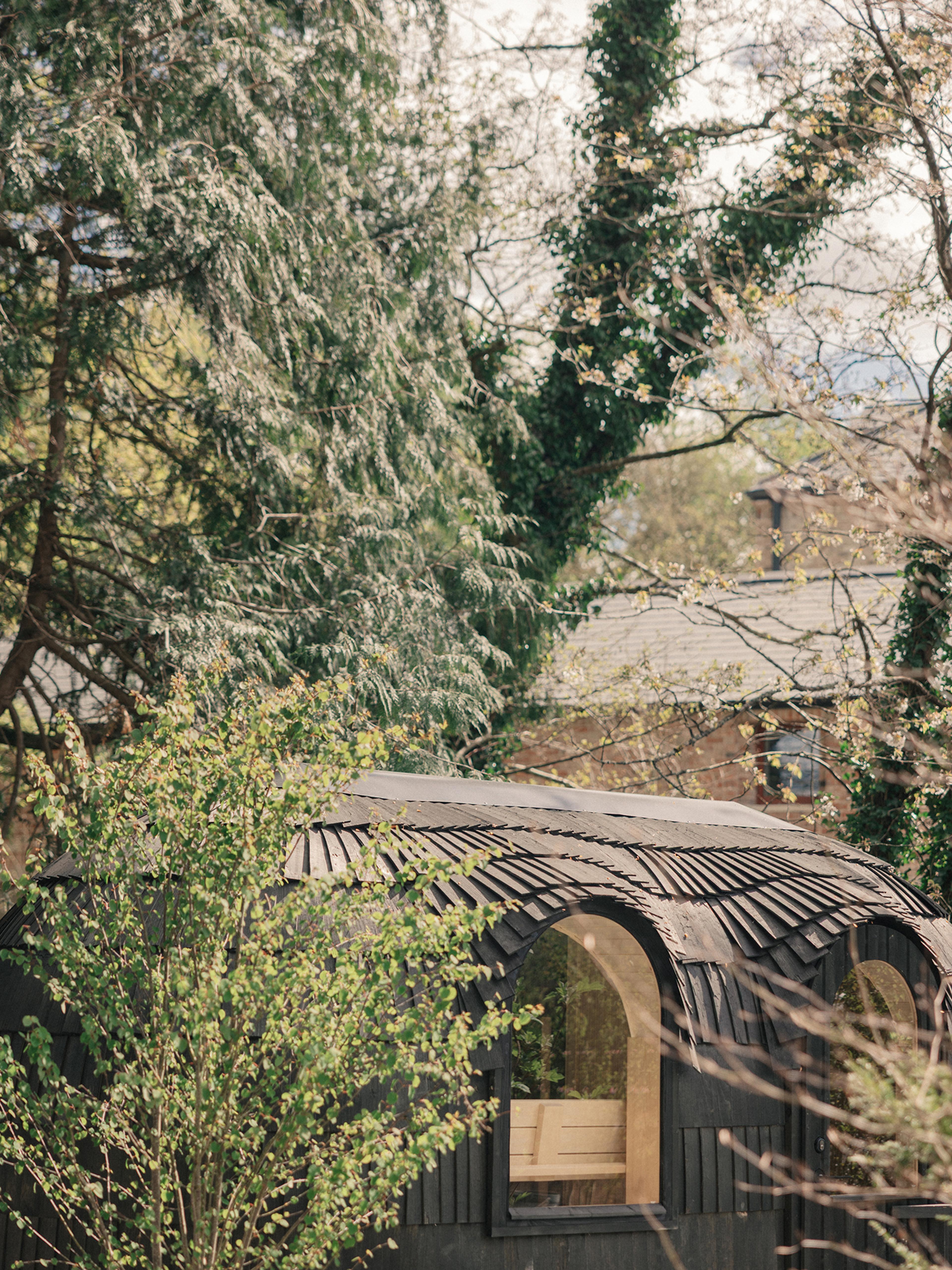An arched, black timber-shingled sauna cabin nestled among tall trees and greenery in the wooded grounds at Ómós