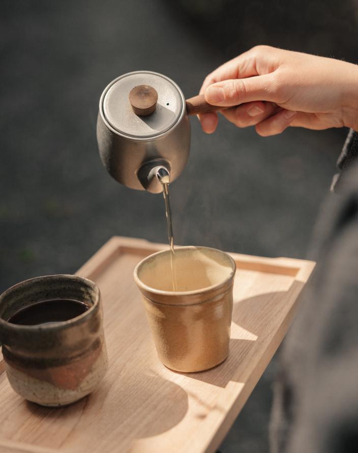 Hand pouring steaming tea from a wood-handled metal teapot into a sandy stoneware cup on a wooden tray, beside a dark glazed cup