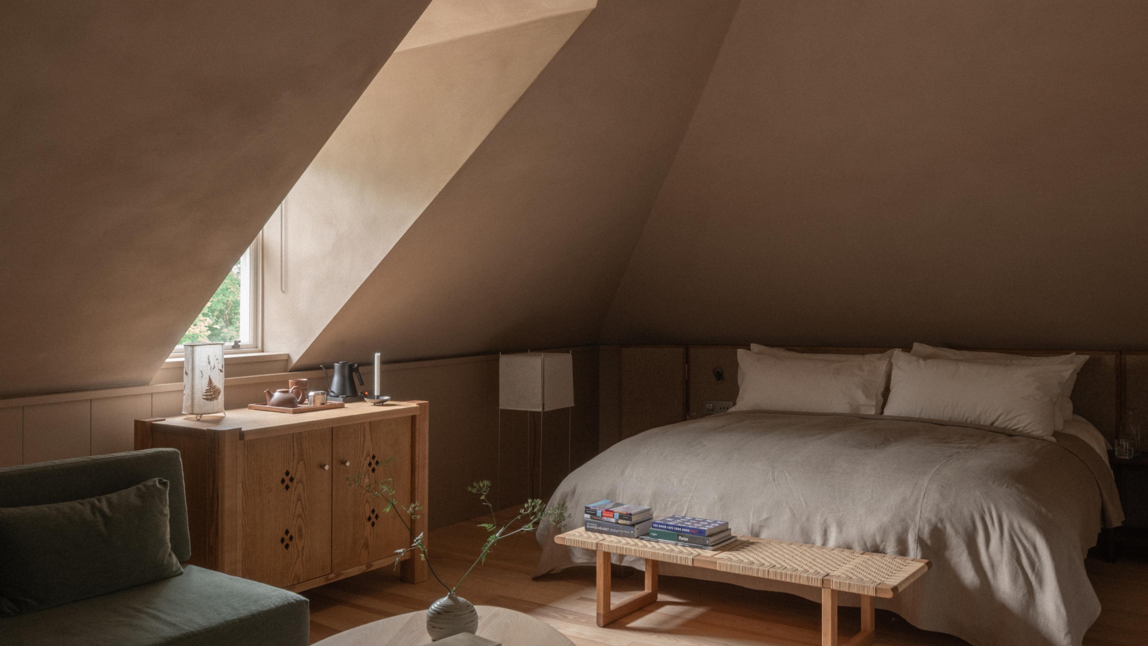 Loft Suite attic bedroom under a sloping plaster ceiling, with a linen-dressed bed, a rattan-topped bench with books, an oak cabinet holding a tea tray and a green sofa