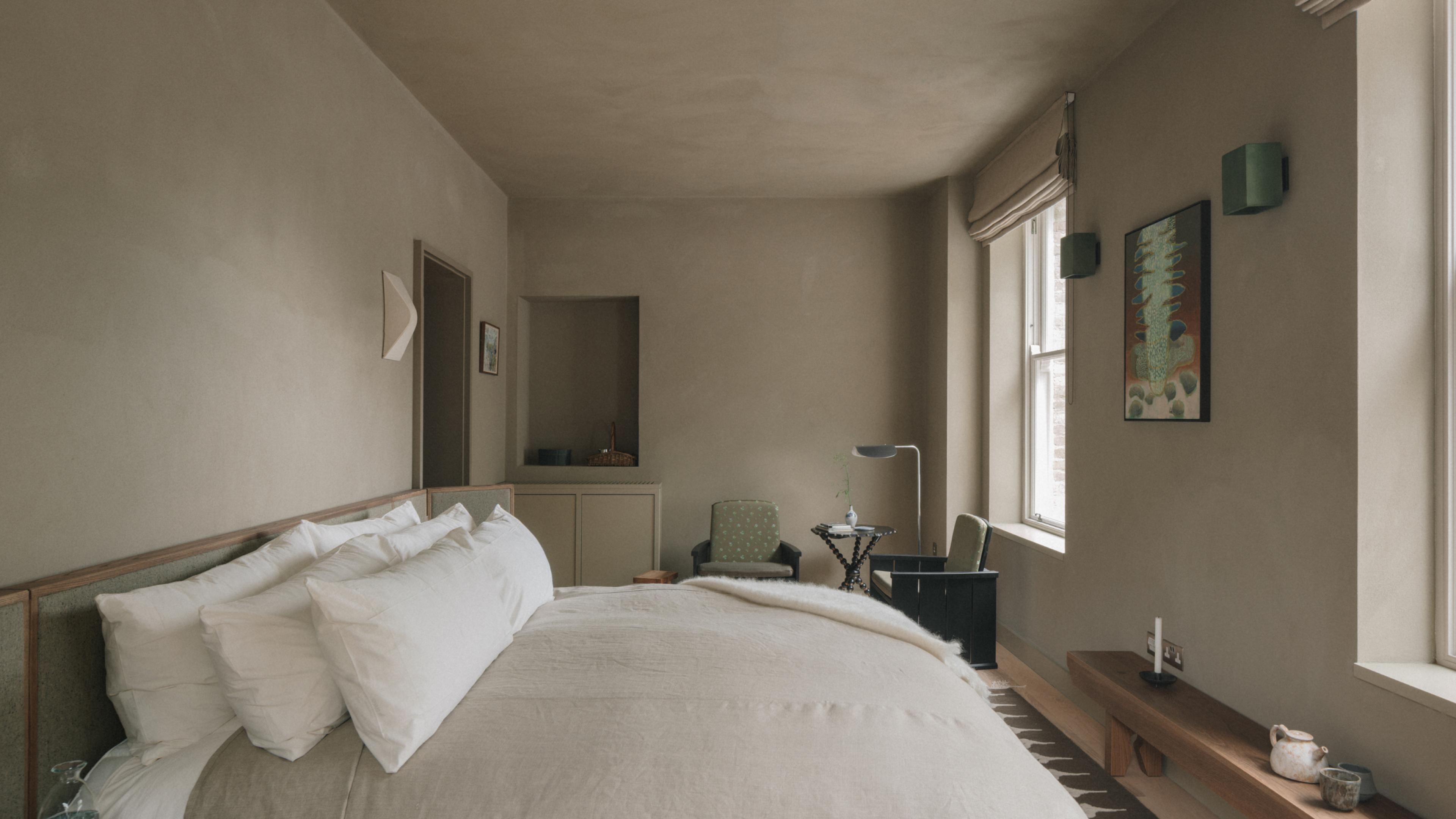 Courtyard View Suite bedroom with a white-linen bed, a reading chair and floor lamp in the corner, a botanical artwork and a low timber bench holding a candlestick and teapot