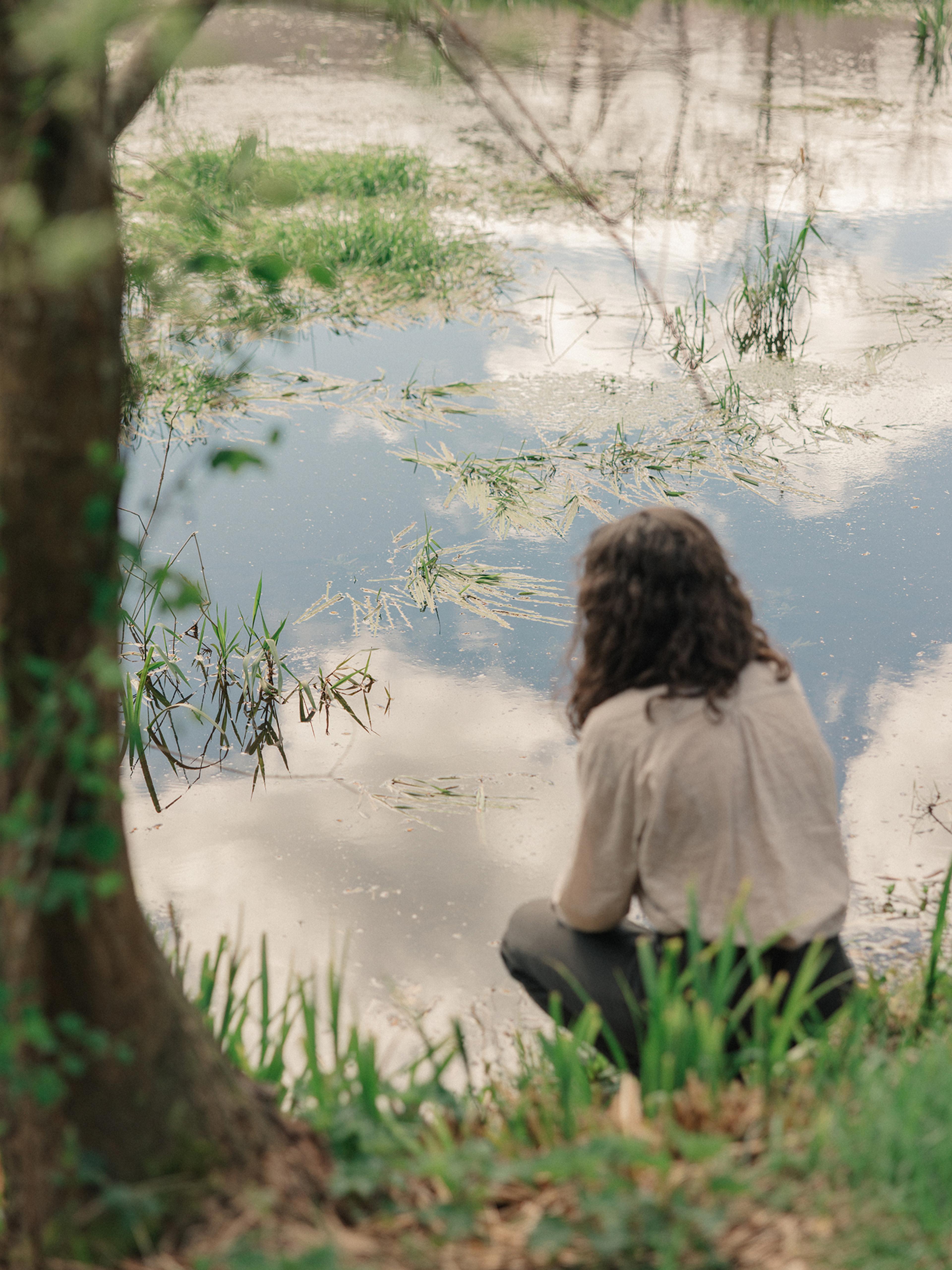 A person in a linen shirt crouches at the edge of a still millpond that mirrors clouds and sky, beside grasses and a tree trunk