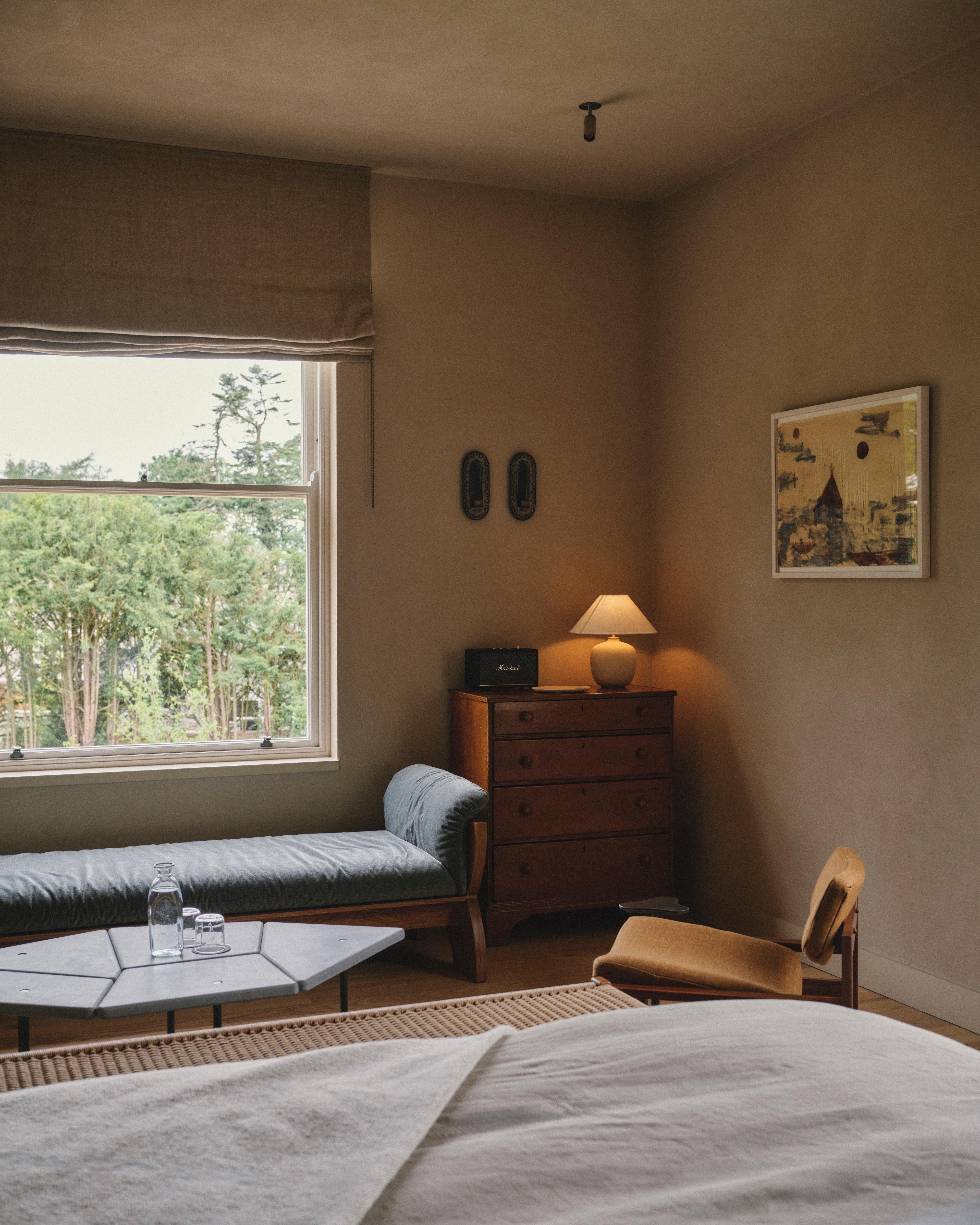 Guest bedroom with a linen-dressed bed, a green daybed beneath a sash window and a wooden chest of drawers with a lamp and framed abstract art