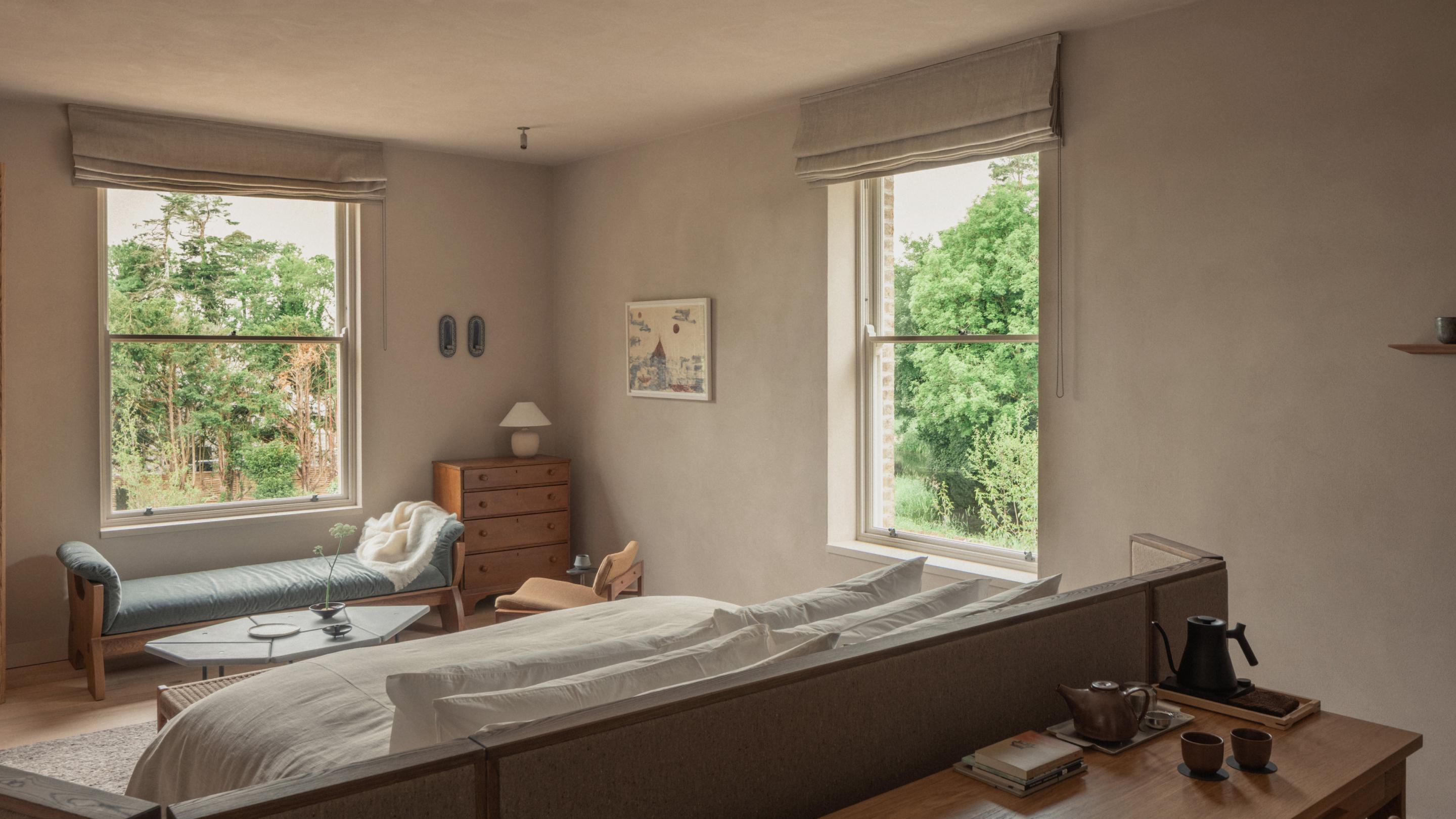 Garden Suite bedroom with a linen-dressed bed, a blue-grey daybed beneath a sash window and a tea tray on the timber console behind the bed, with leafy views outside