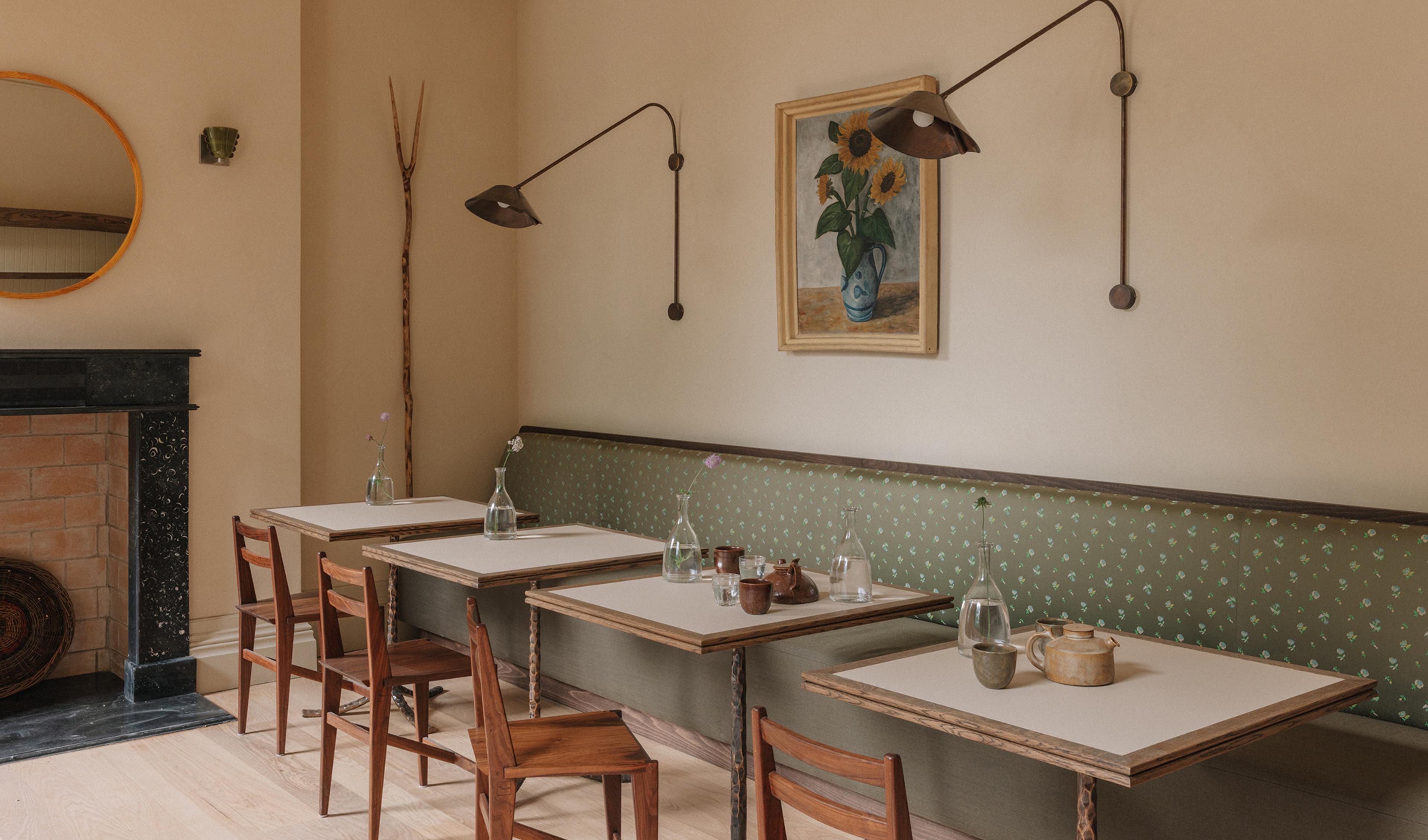 Breakfast room with wooden chairs at pale-topped tables along a green sprigged banquette, dark swing-arm wall lights flanking a framed sunflower painting, and a black marble fireplace in the corner