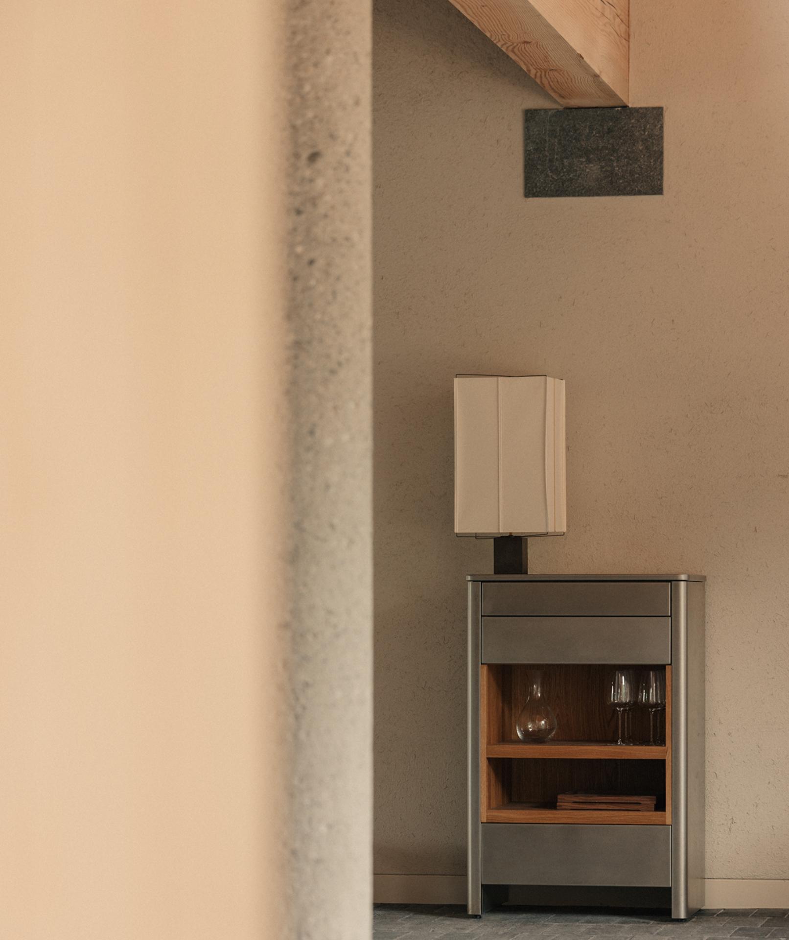 A metal and oak cabinet holding a glass decanter and stemware, lit by a shaded lamp against a plaster wall