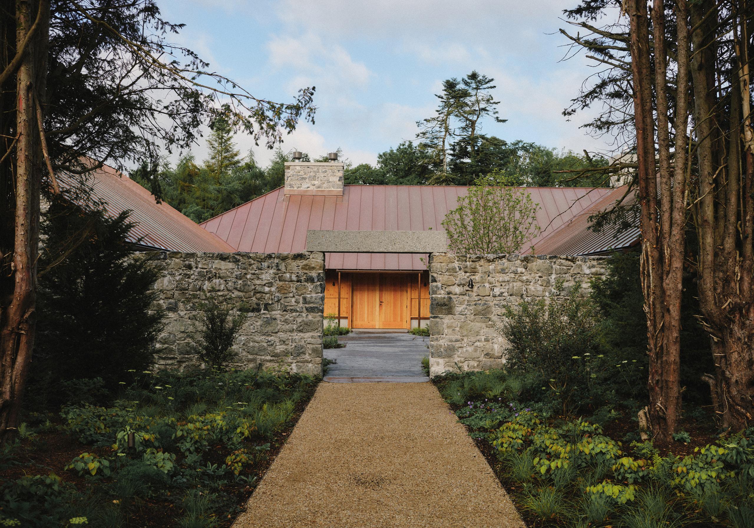Stone restaurant building with copper roof and timber doors framed by tall trees, gravel path leading to the entrance
