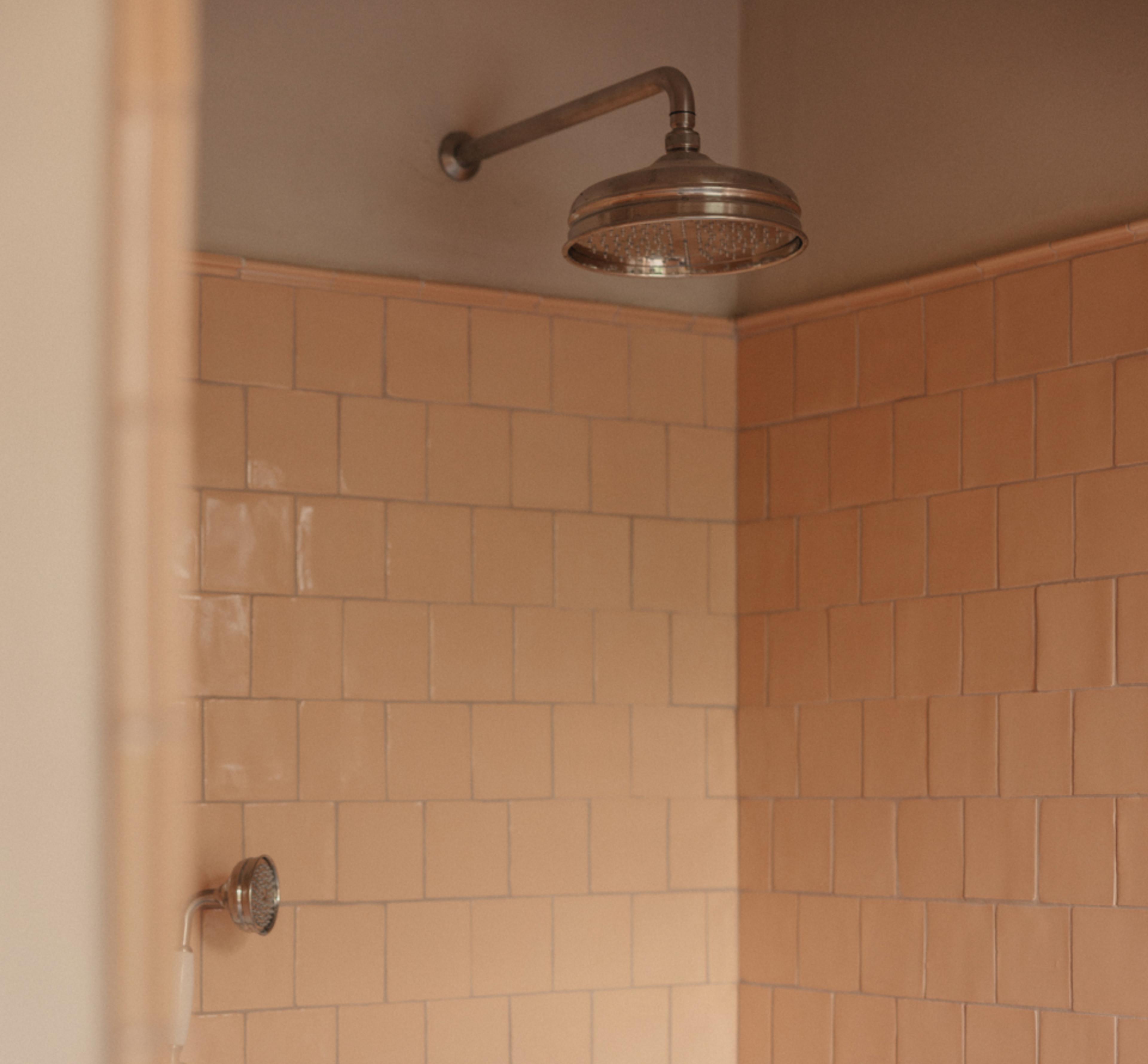 Large vintage-style nickel rain showerhead on a corner wall of warm peach-toned square tiles, with a smaller handheld shower fitting below