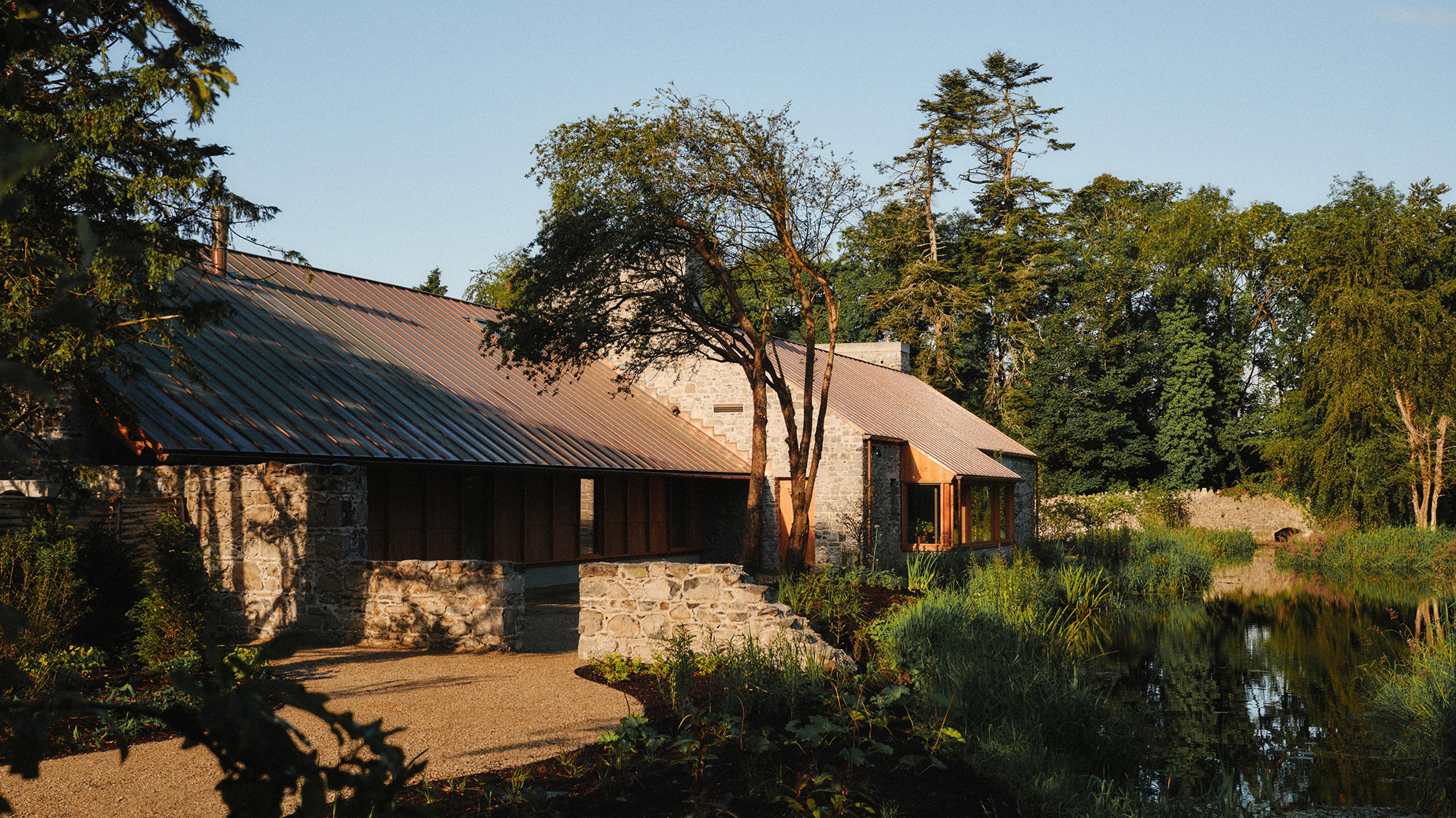 Stone restaurant with metal roof beside a still millpond in warm evening light, surrounded by trees and greenery