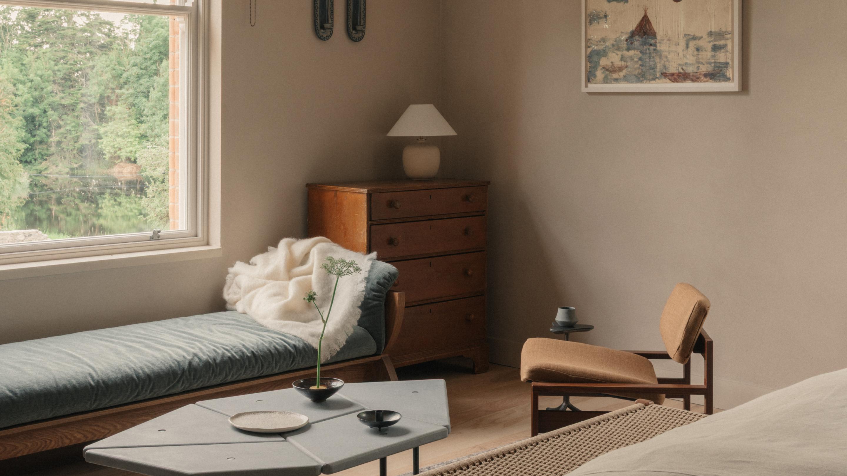 Garden Suite sitting corner with a blue-grey daybed and white throw by a window overlooking trees and water, a wooden chest of drawers with a lamp, a tan lounge chair and a stone-topped coffee table