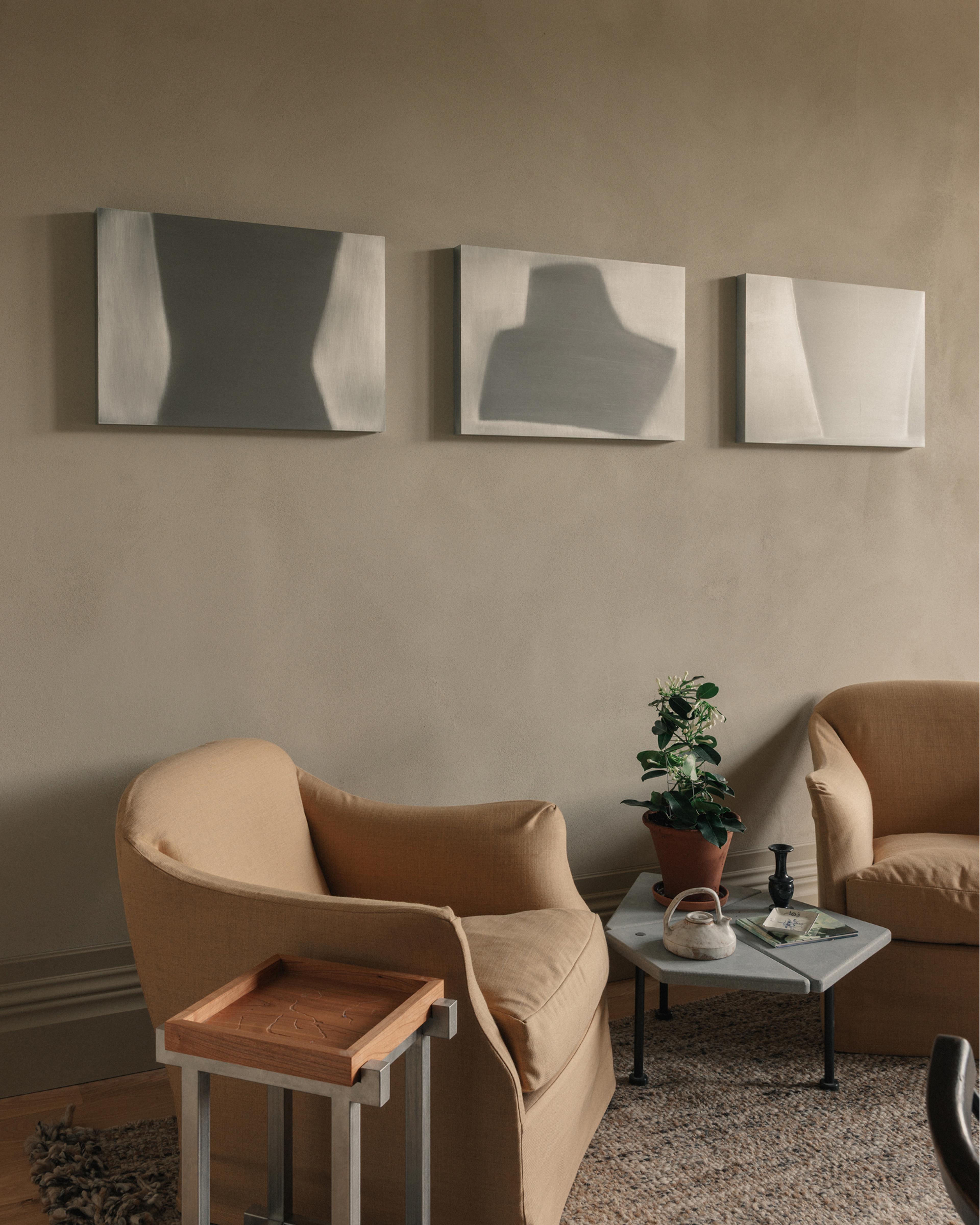 Sitting room with three soft grey abstract canvases above two tan armchairs, a small stone table with a potted plant and teapot on a woven rug