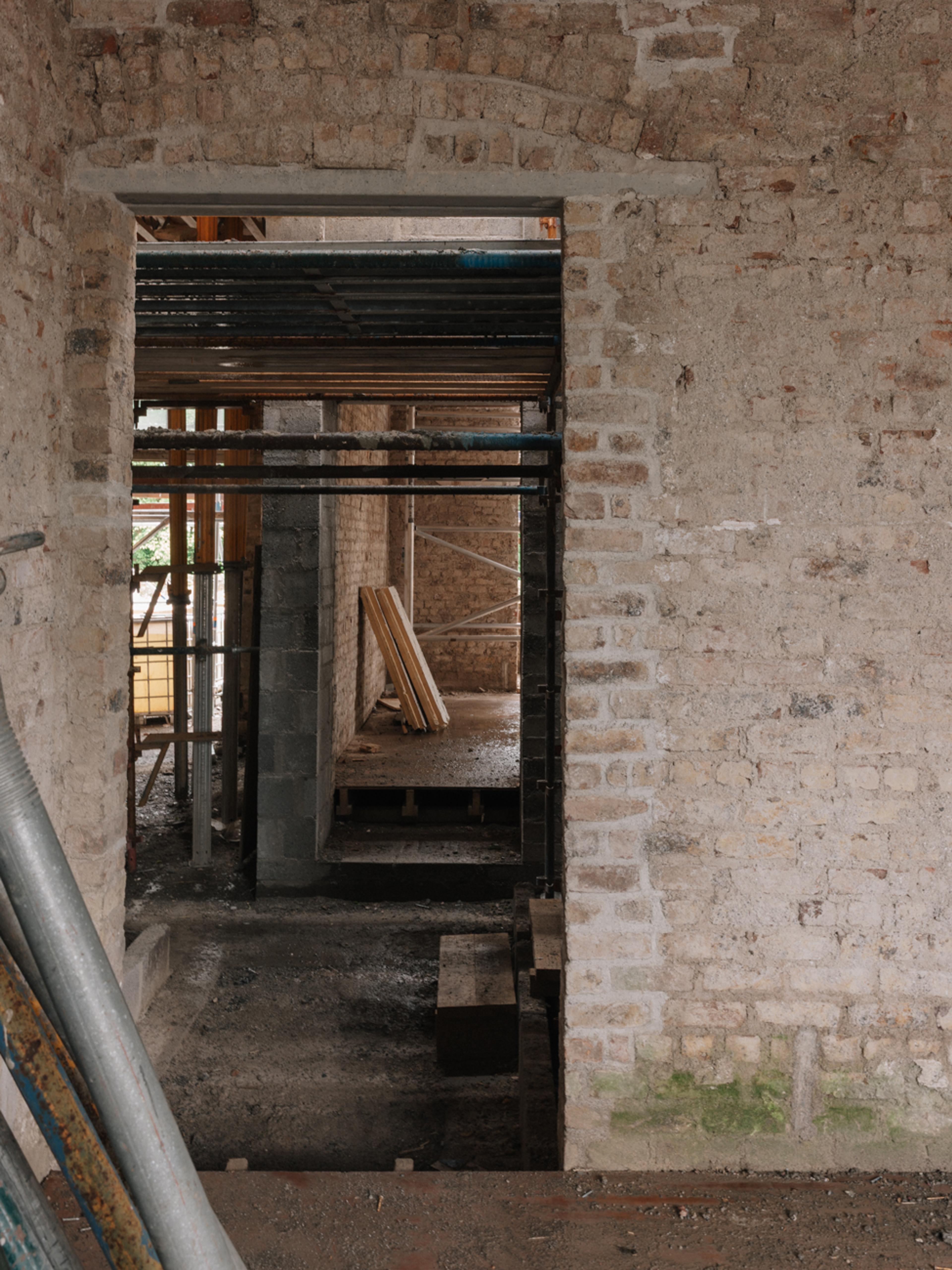 View through a doorway in an exposed bare-brick room under renovation, revealing scaffolding poles, timber boards, and a concrete block partition wall inside