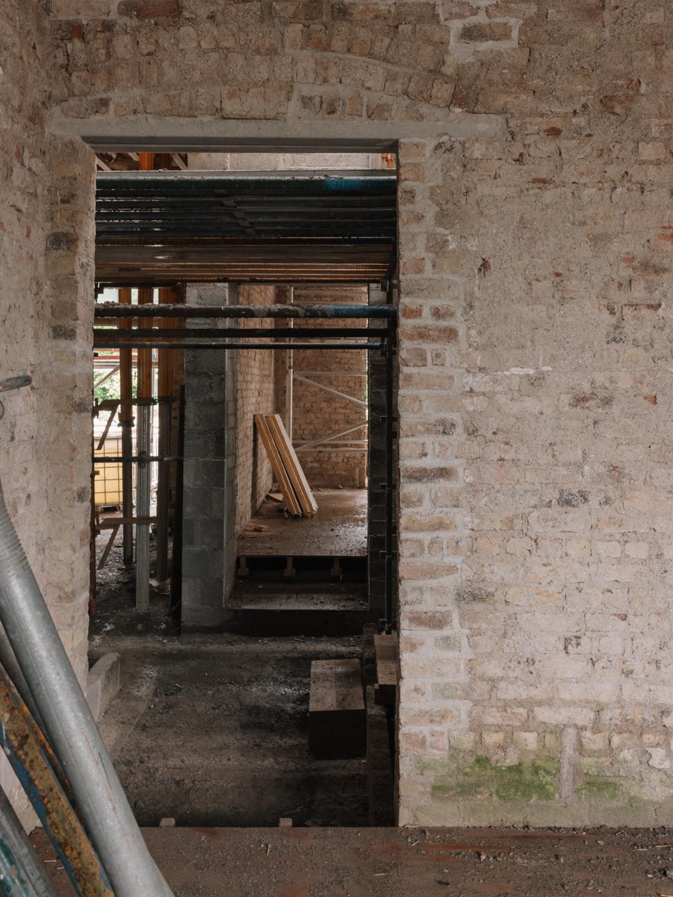 View through a doorway in an exposed bare-brick room under renovation, revealing scaffolding poles, timber boards, and a concrete block partition wall inside