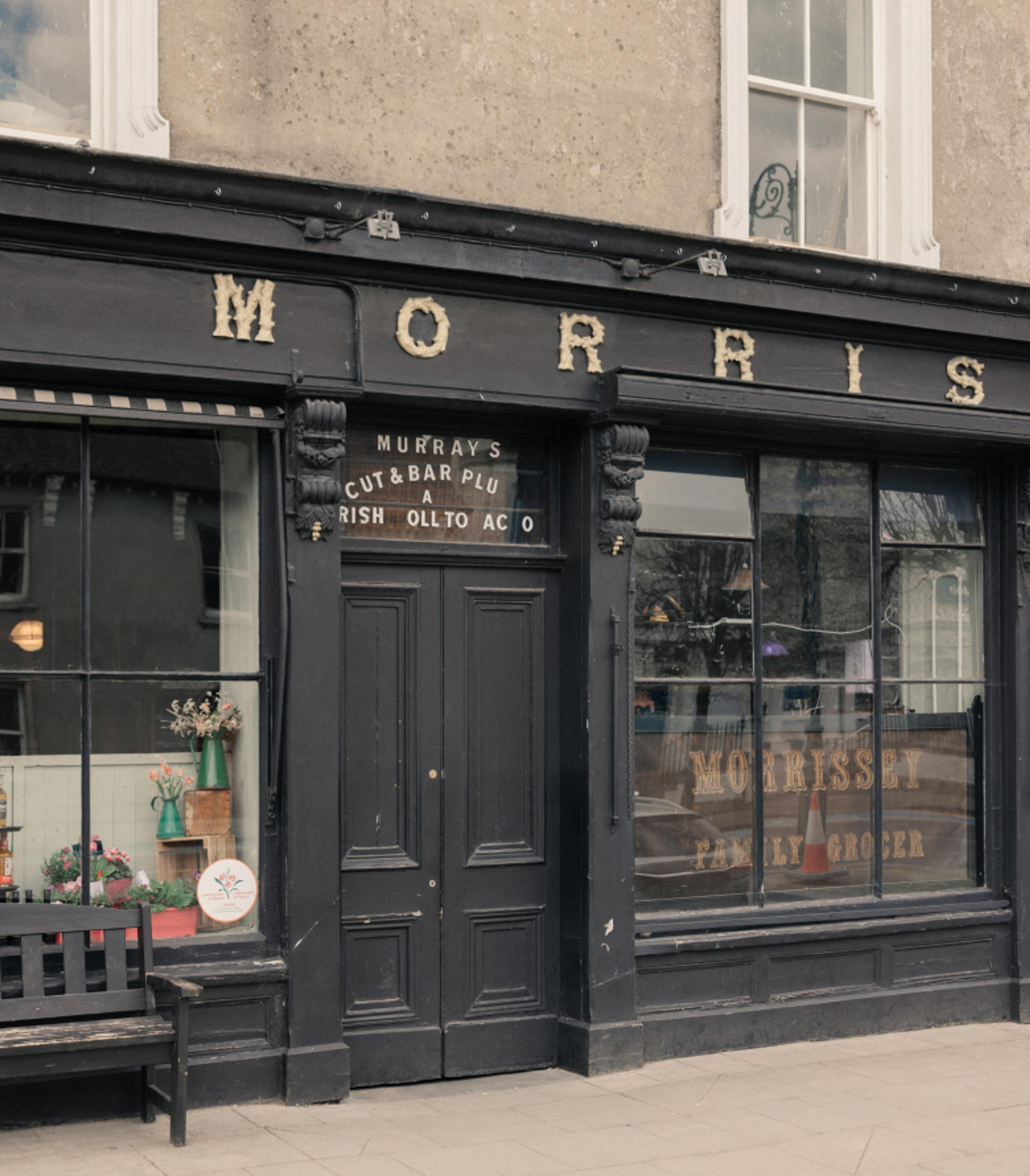 The black timber shopfront of Morrissey's family grocer and pub in Abbeyleix, with gilded lettering, a bench and a flower-filled window