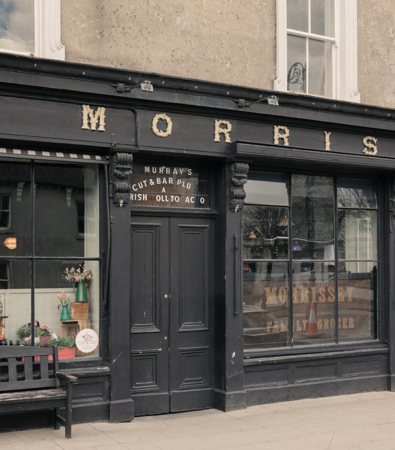 The black timber shopfront of Morrissey's family grocer and pub in Abbeyleix, with gilded lettering, a bench and a flower-filled window