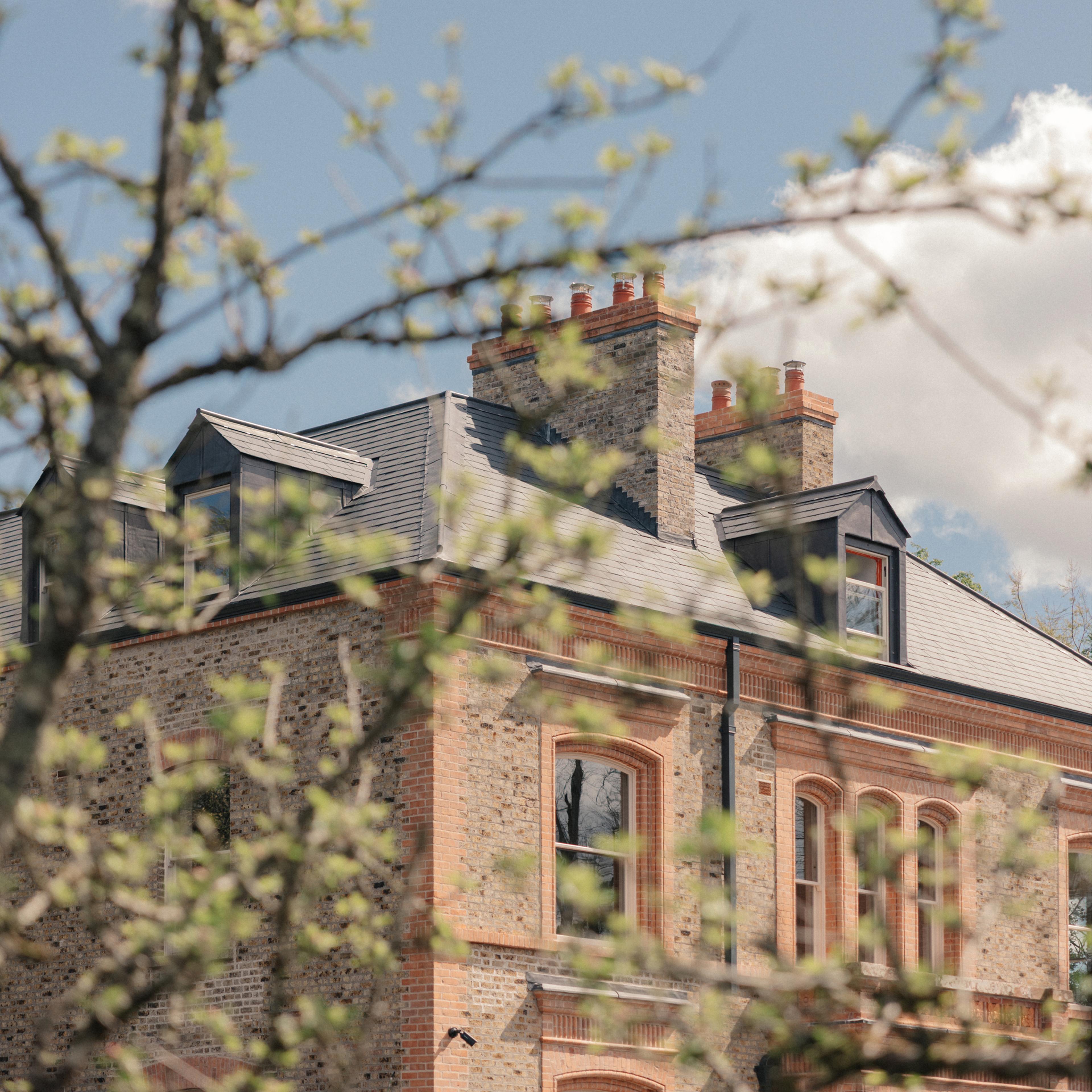 Red-brick and stone guesthouse with slate dormer roofs and chimneys, glimpsed through the budding spring branches of a tree under a blue sky