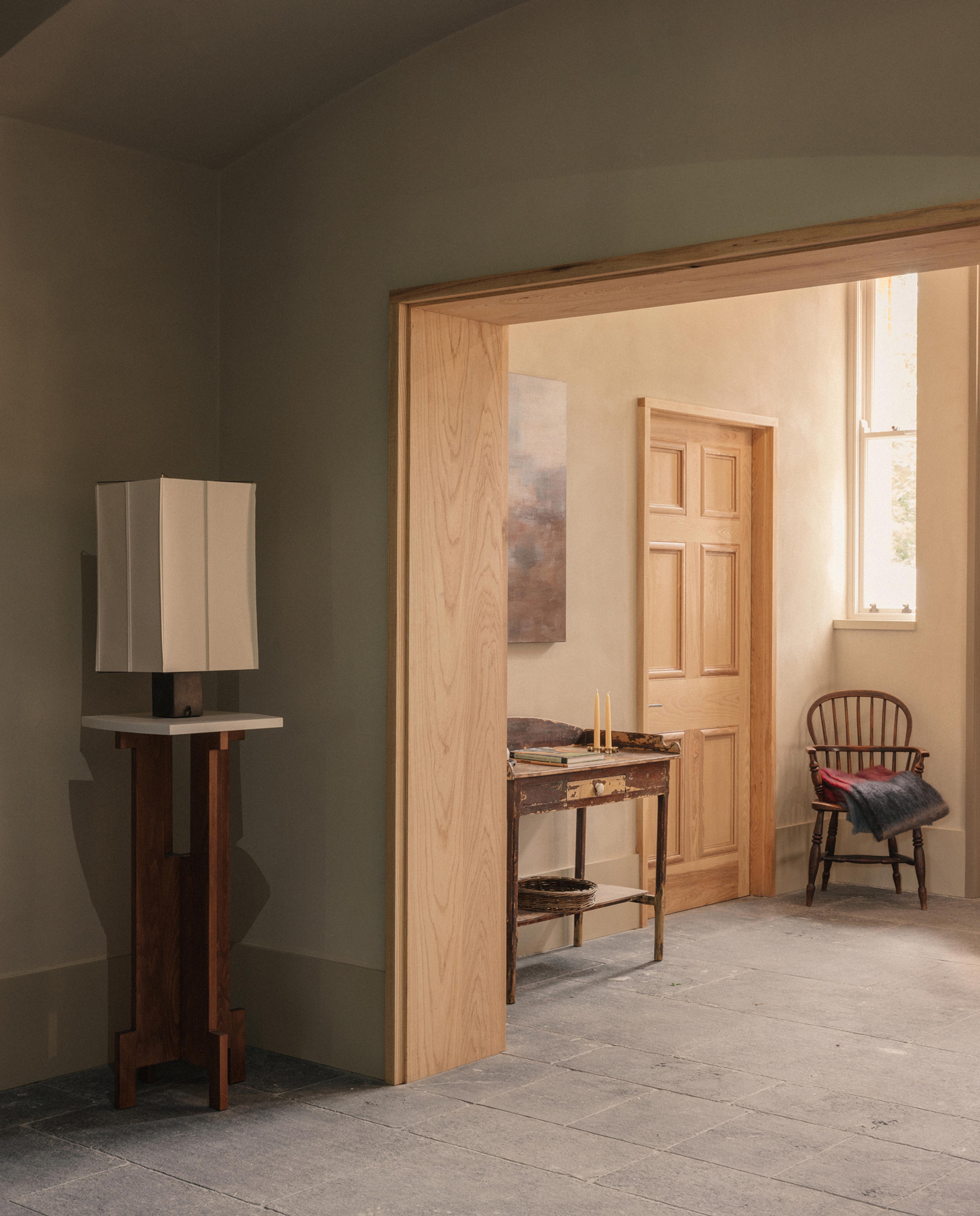 Sage-green room with a square table lamp on a wooden plinth, looking through a timber doorway to a rustic console table with candles and a spindle-back chair