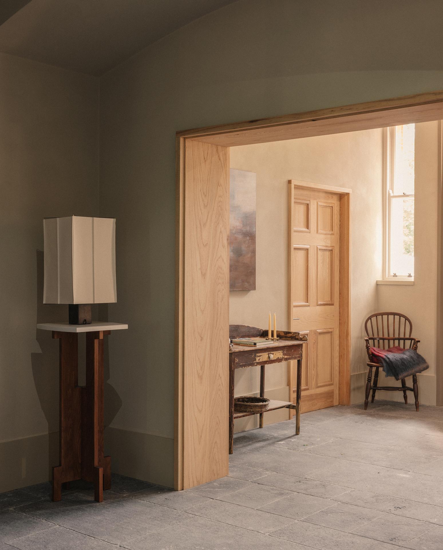 Sage-green room with a square table lamp on a wooden plinth, looking through a timber doorway to a rustic console table with candles and a spindle-back chair