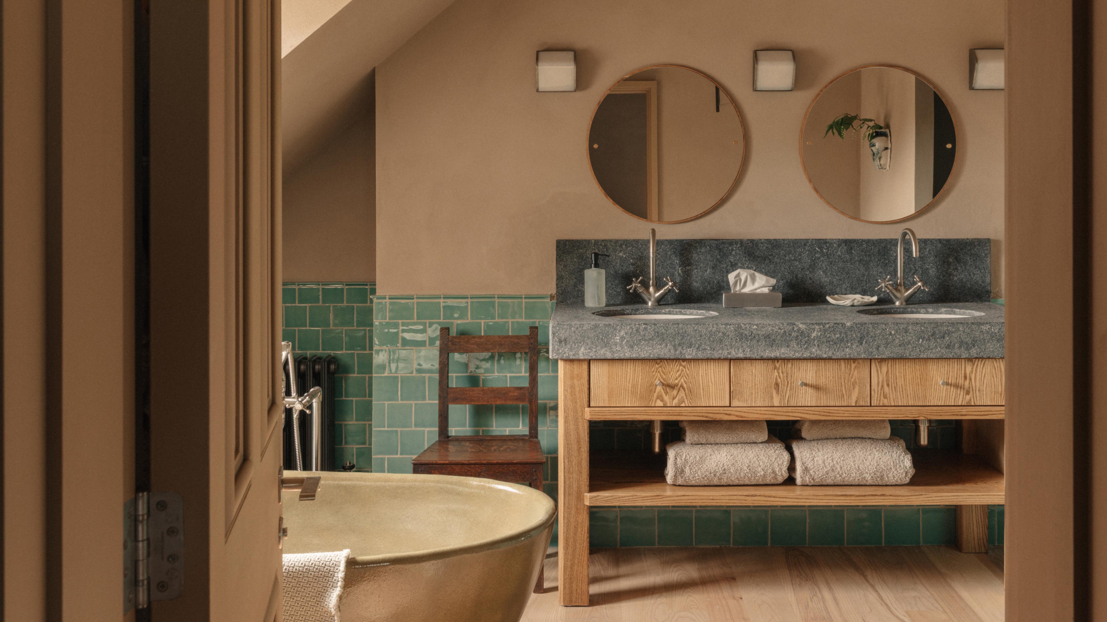 Loft Suite bathroom with a brass freestanding bath, green-tiled walls, a dark wooden chair and a stone-topped double ash vanity beneath two round mirrors