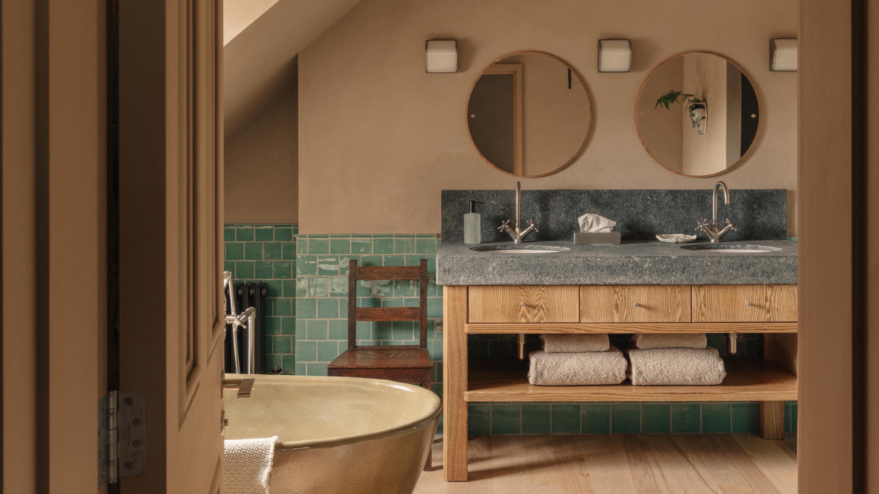 Loft Suite bathroom with a brass freestanding bath, green-tiled walls, a dark wooden chair and a stone-topped double ash vanity beneath two round mirrors