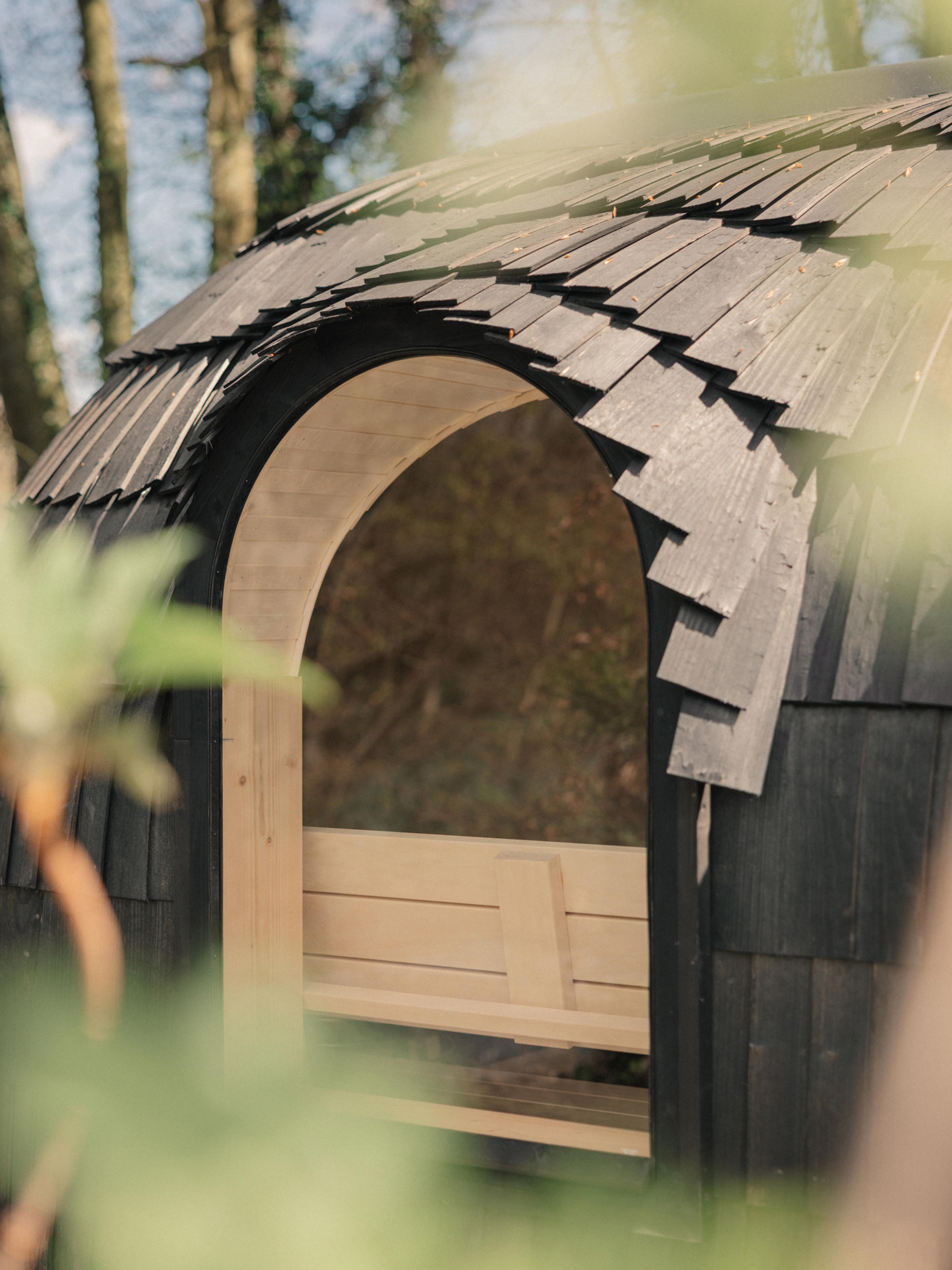 Close-up of the arched window and overlapping black shingles of the Ómós outdoor sauna, with a pale wood bench glimpsed inside