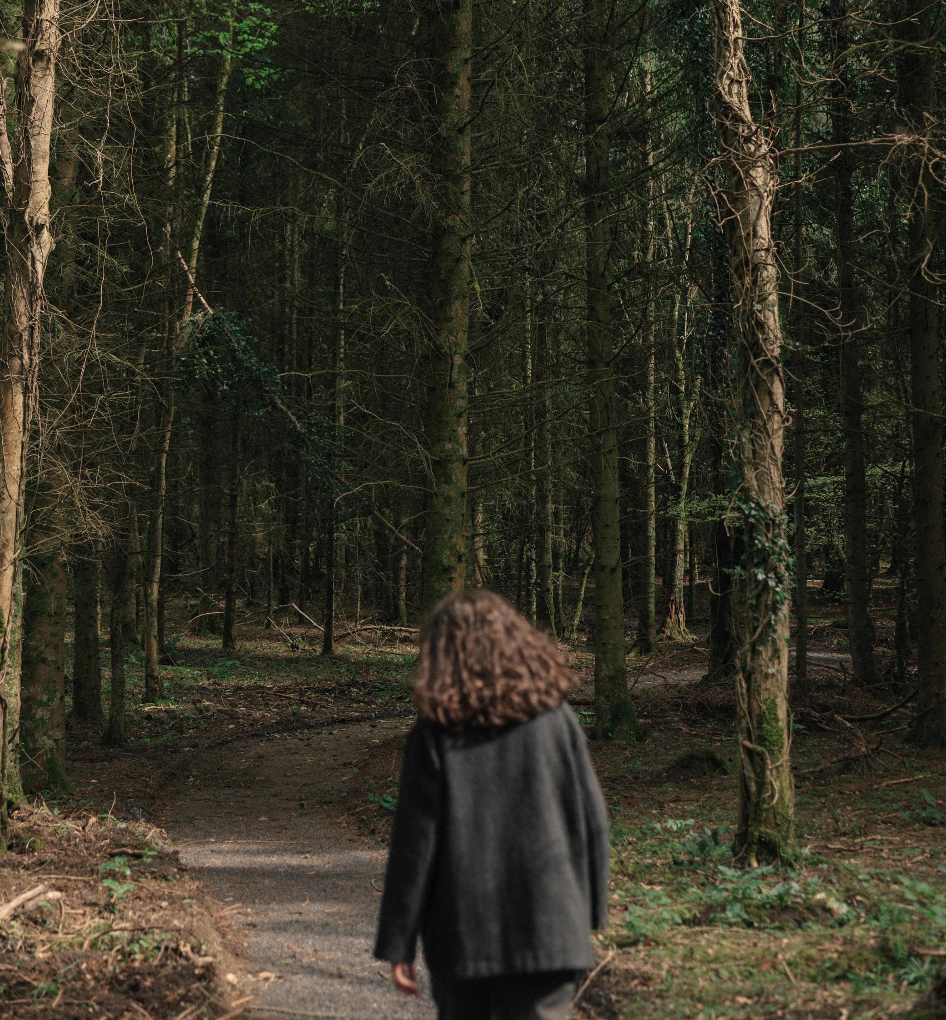 A person walks alone along a winding path through a dense, shadowy forest of tall conifers near Ómós