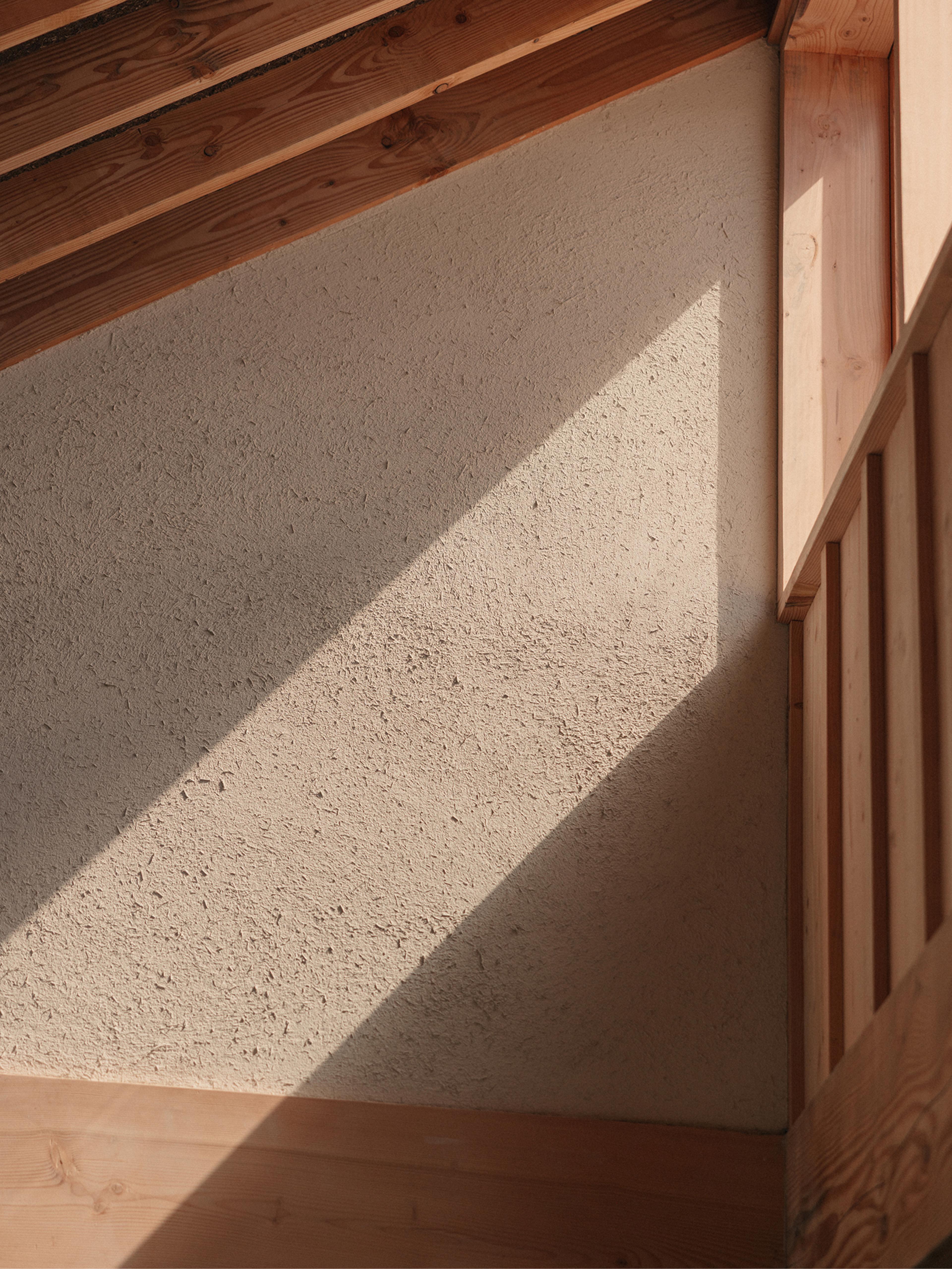 A diagonal shaft of sunlight falling across a textured plaster wall framed by warm pine timber beams and panelling at Ómós
