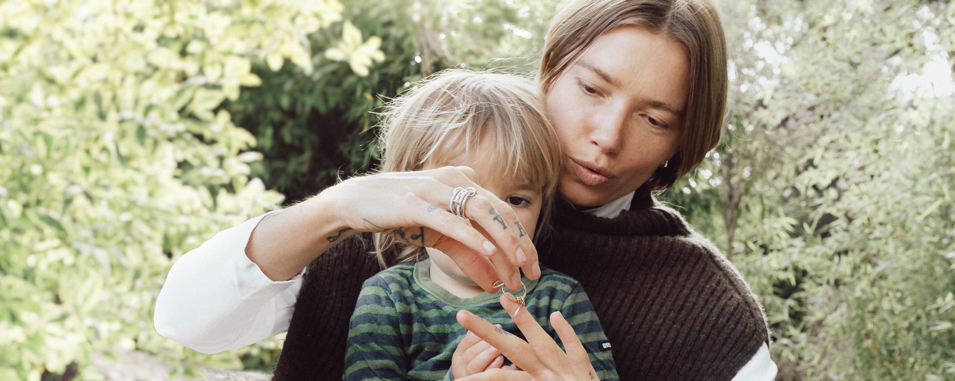 Spinelli Kilcollin mother and child sit in lush landscape with a child on a mother's lap showing the child a Spinelli Kilcollin ring partly on her finger