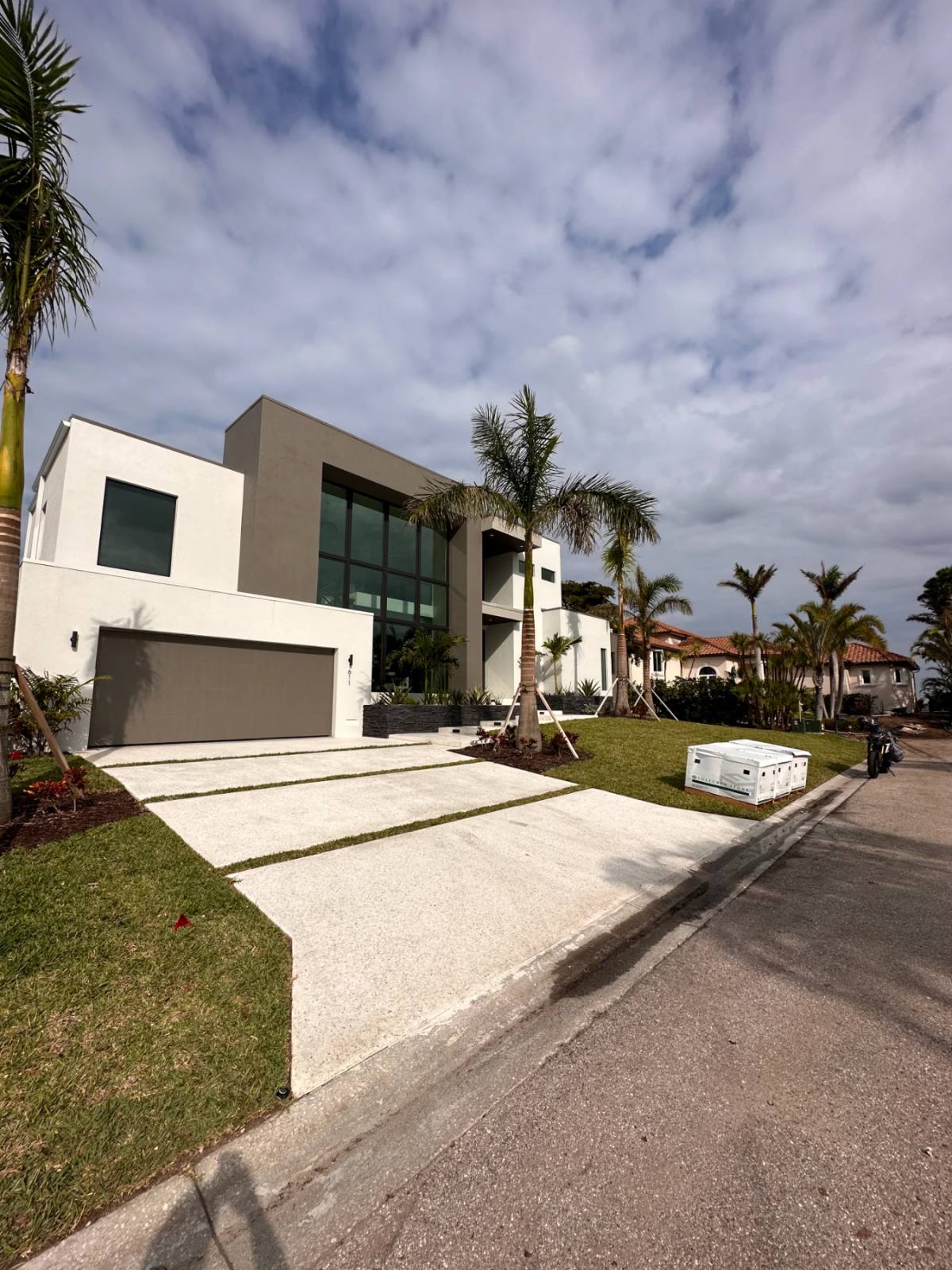 A wide pale driveway in long panels with grass joints running from the street up to a modern two-storey house with a grey garage door, palms along the curb