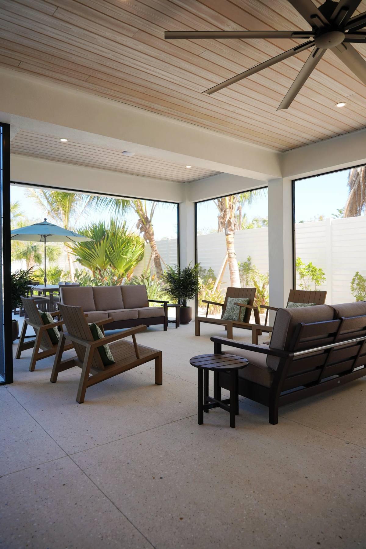 A covered lanai on a coastal build — outdoor seating on a pale speckled floor, palms beyond the screen