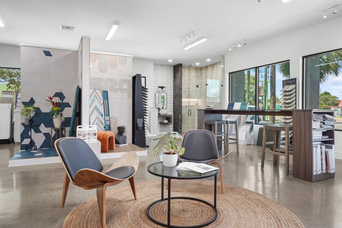 Tile showroom lounge with two walnut shell chairs on a round jute rug facing a blue geometric tile feature wall and a bathroom vignette