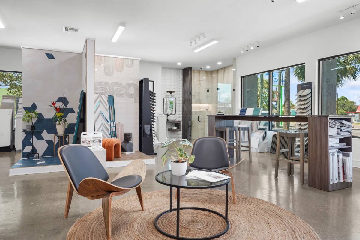 Tile showroom lounge with two walnut shell chairs on a round jute rug facing a blue geometric tile feature wall and a bathroom vignette