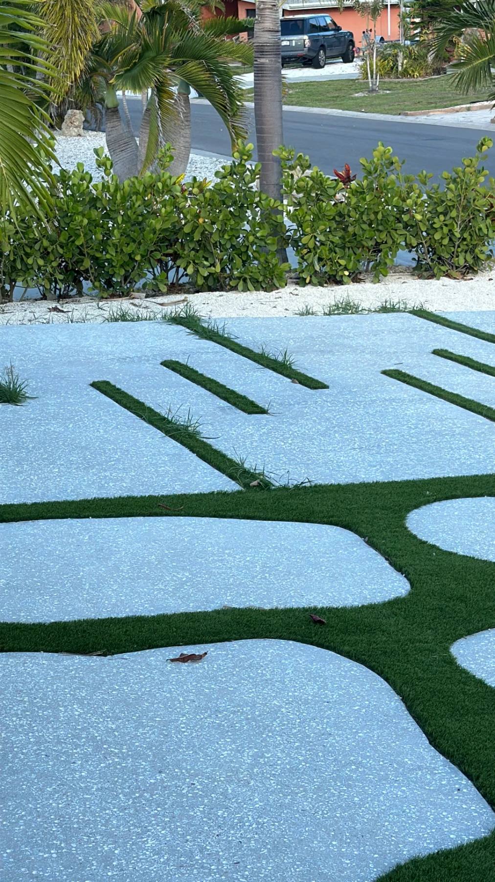 A pale driveway of narrow turf slots and large freeform islands edged in turf, running out to a hedge and the street