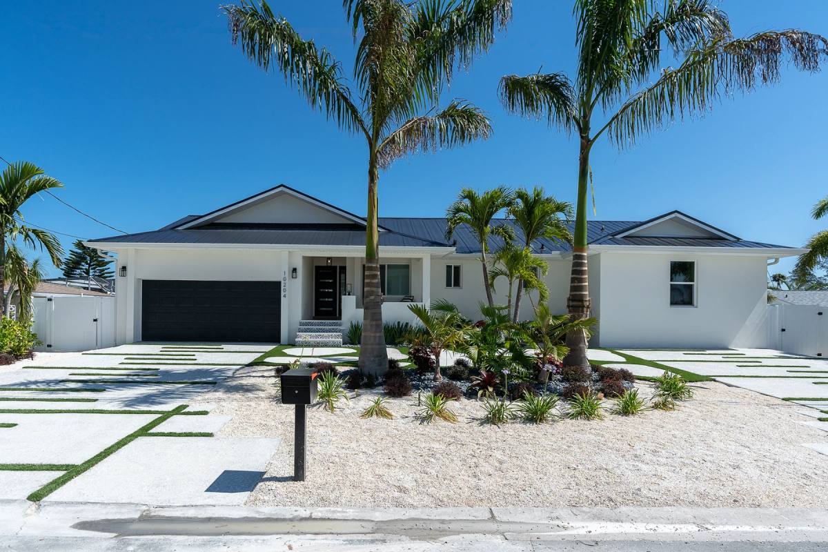 Straight-on view of a white single-storey house with a metal roof, a pale driveway of slabs and turf spreading across the frontage, palms and a black mailbox