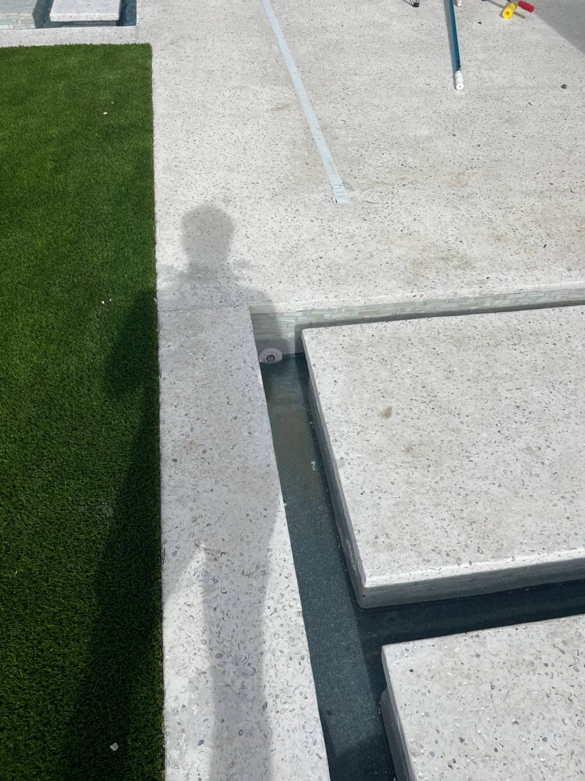 Overhead view of pale shell-aggregate deck slabs meeting artificial turf on one side and a narrow water channel on the other