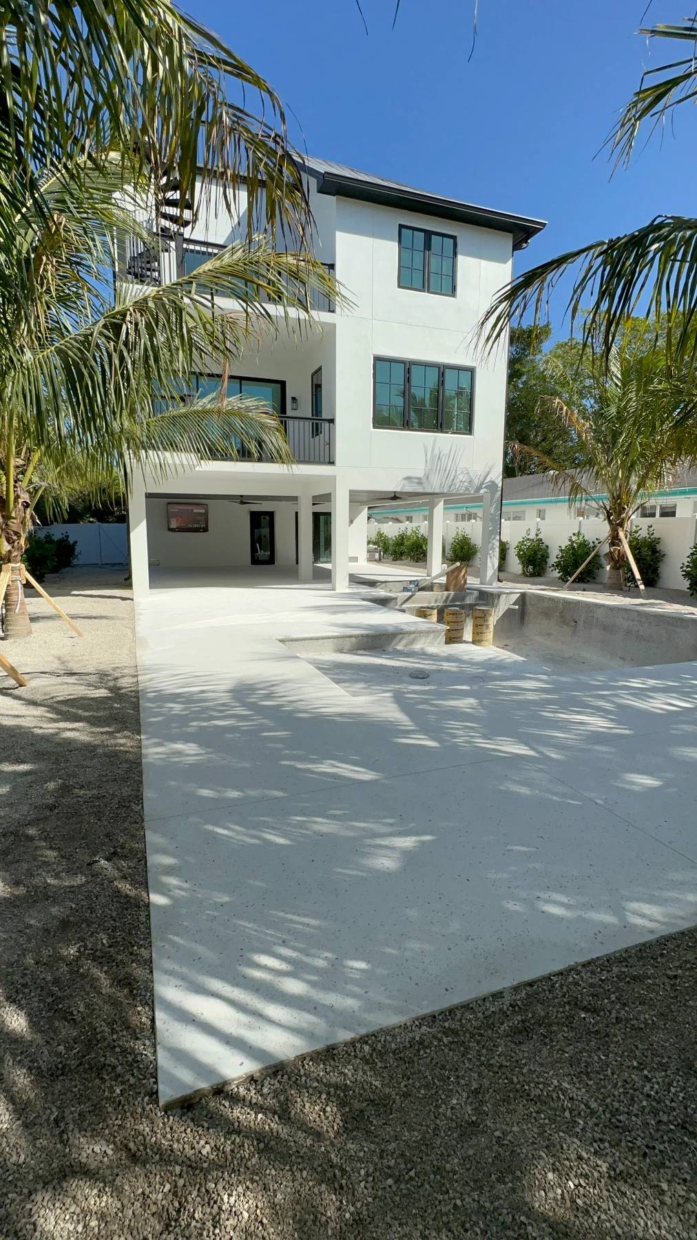 Freshly poured pale deck, not yet ground, wrapped around an empty pool shell beside the open ground floor of a raised white house, with newly staked palms in the sand