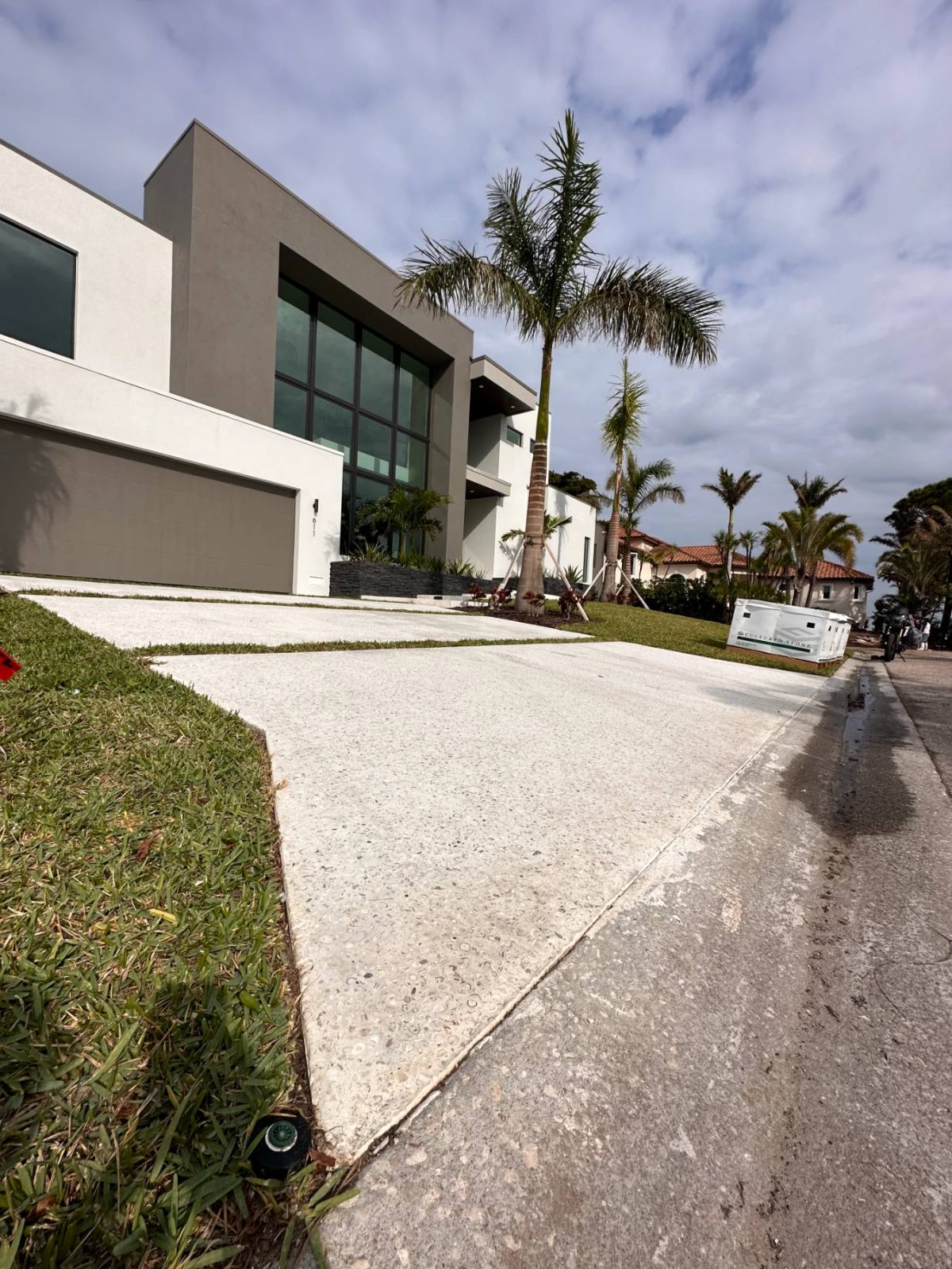 Low view down a long pale driveway to the street, a lawn along one side and a modern grey and white house with palms behind