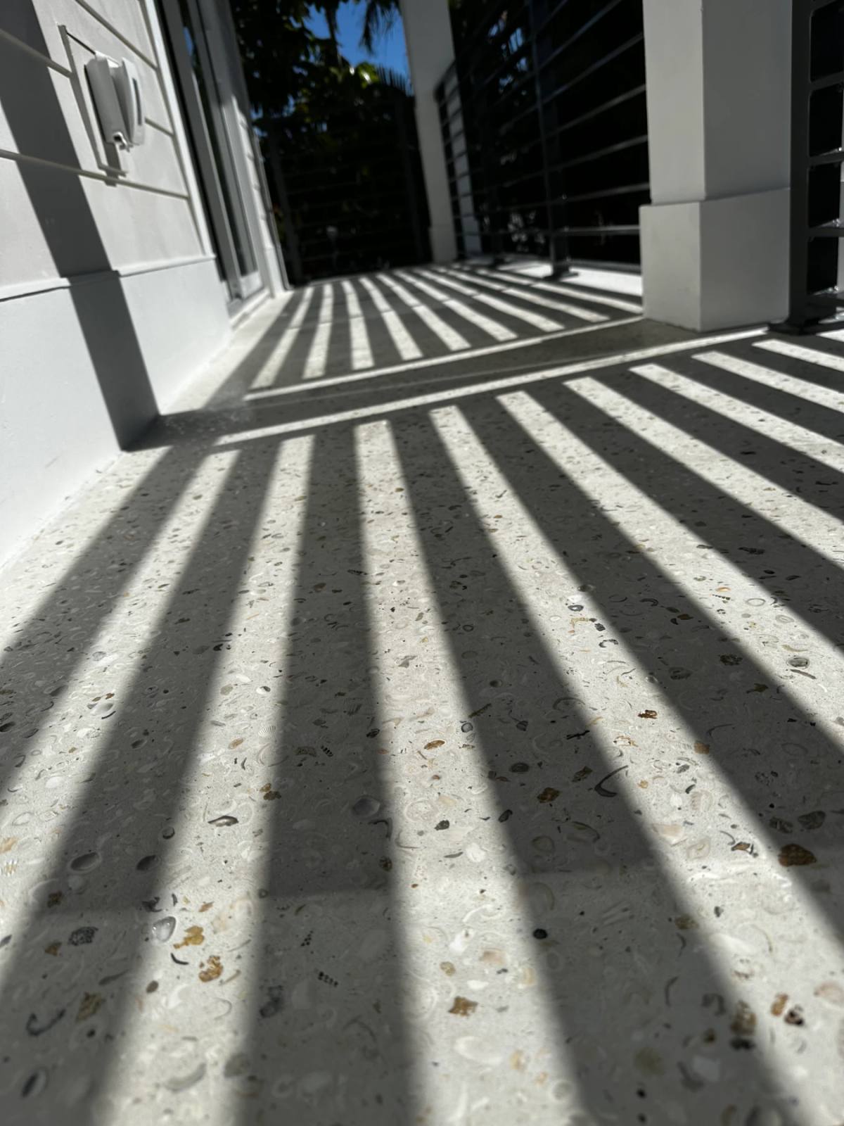 Shadows of a balcony railing falling in long stripes across a pale Shellcrete terrace floor, shell pieces visible in the sunlit bands