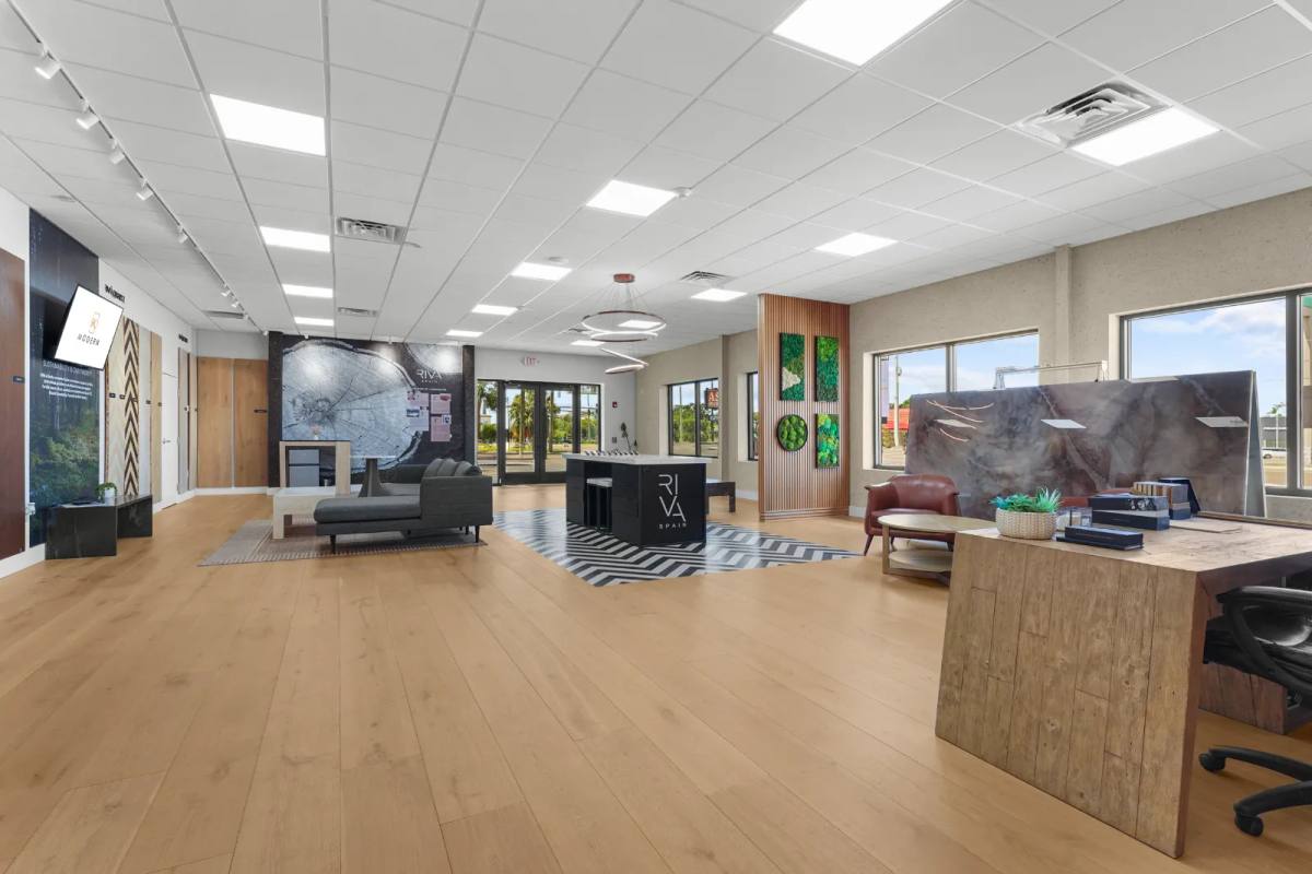 A wide flooring showroom with oak plank floors, a sample island, lounge seating and full-height windows toward the glass entry doors
