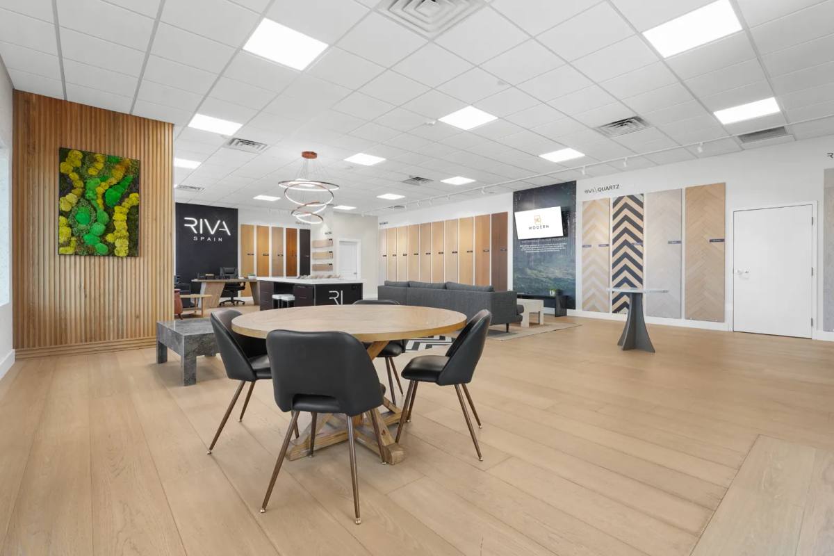 A flooring showroom with a round oak meeting table and black chairs, framed moss art on a slatted wood column and plank sample walls beyond